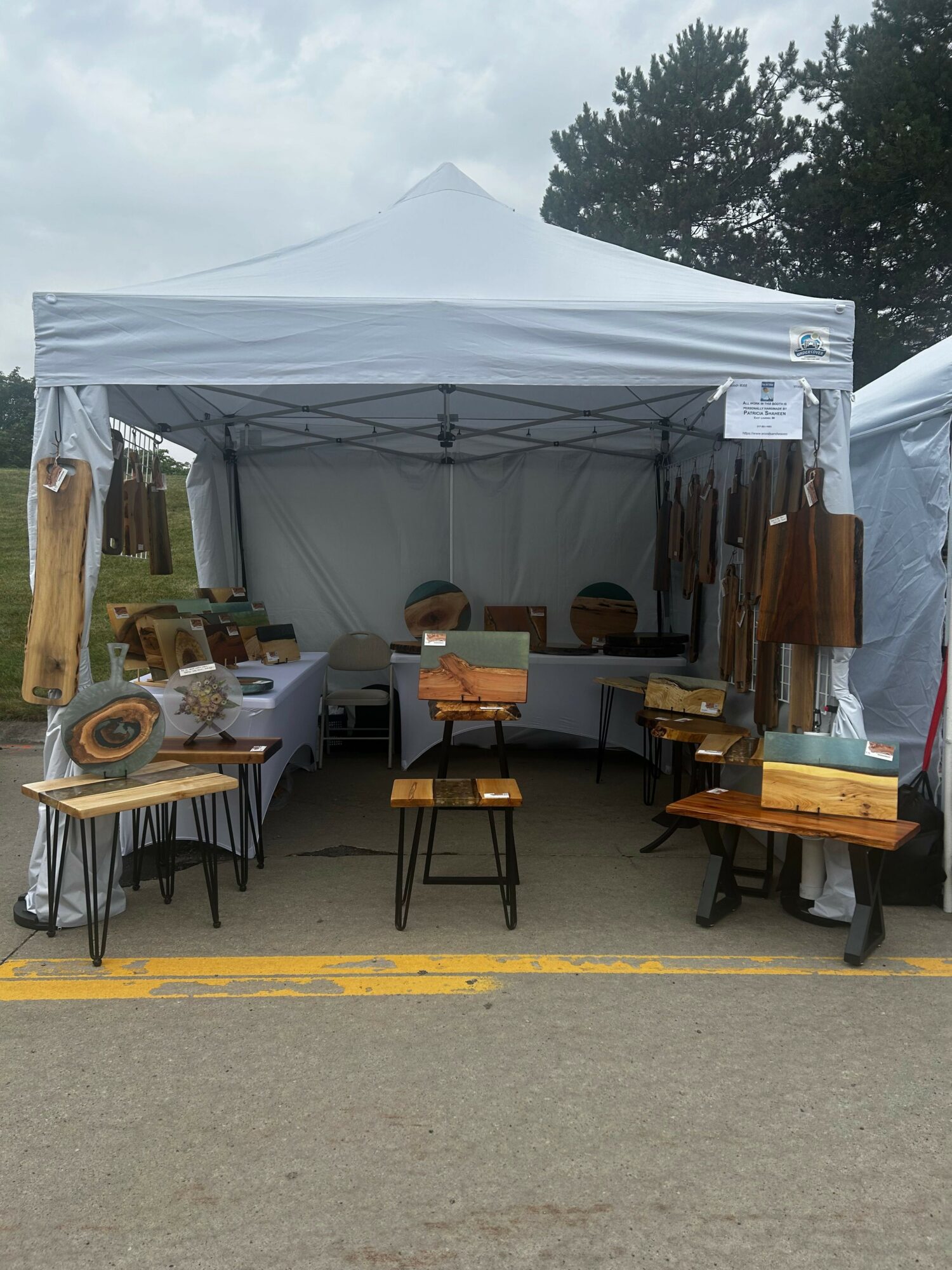 Outdoor booth with wooden items on tables and chairs under a white canopy, on a paved surface with trees in background.