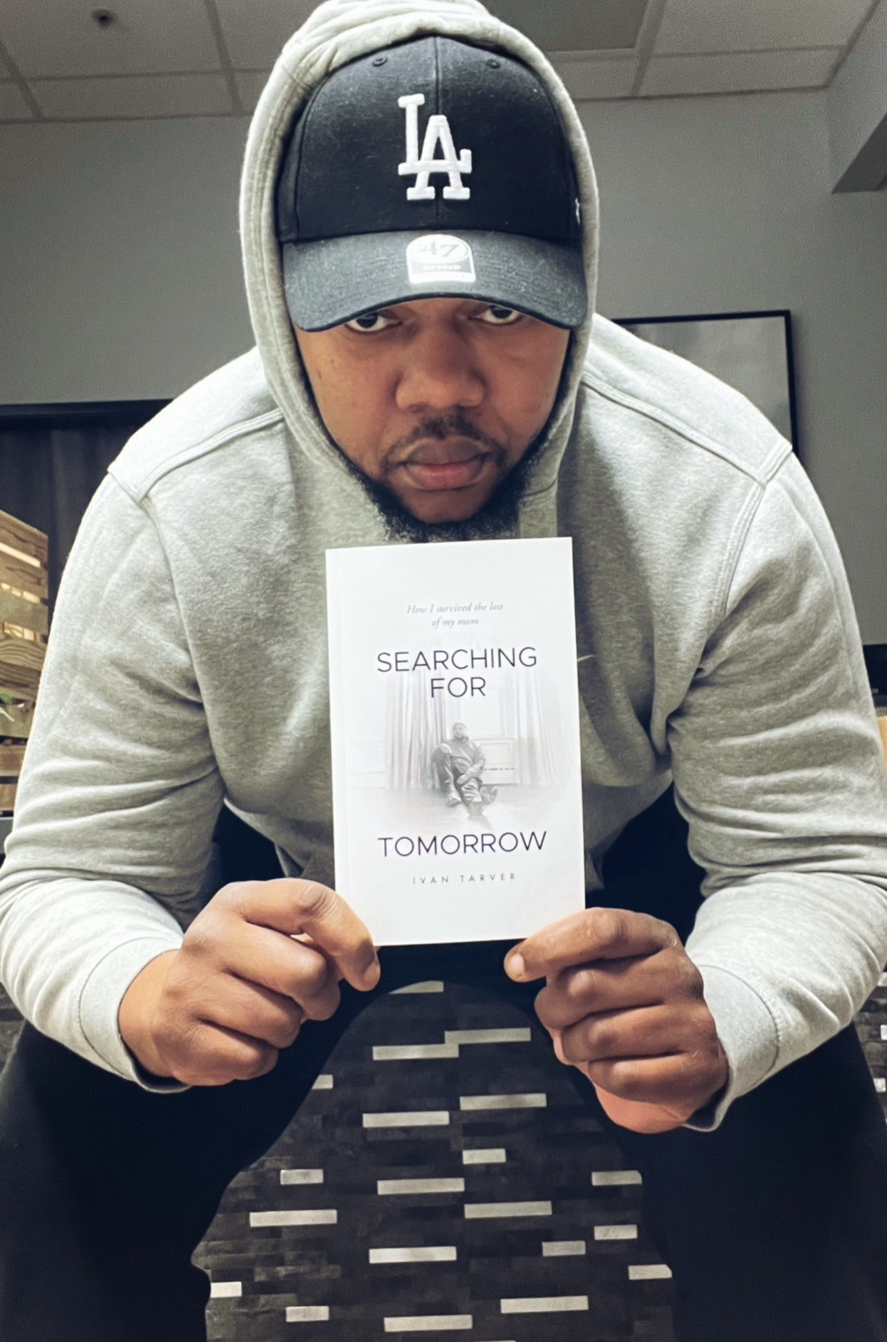 Man in hoodie and Los Angeles cap holding a book titled 'Searching for Tomorrow' indoors.