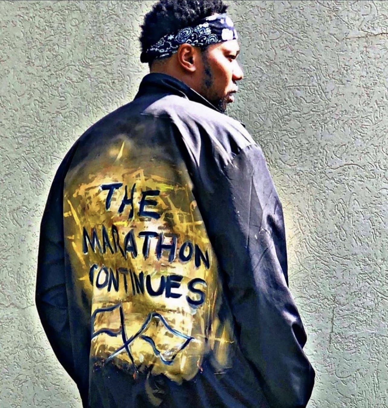 Person with a bandana facing sideways, wearing a jacket with painted text and a landscape, standing against a textured wall.