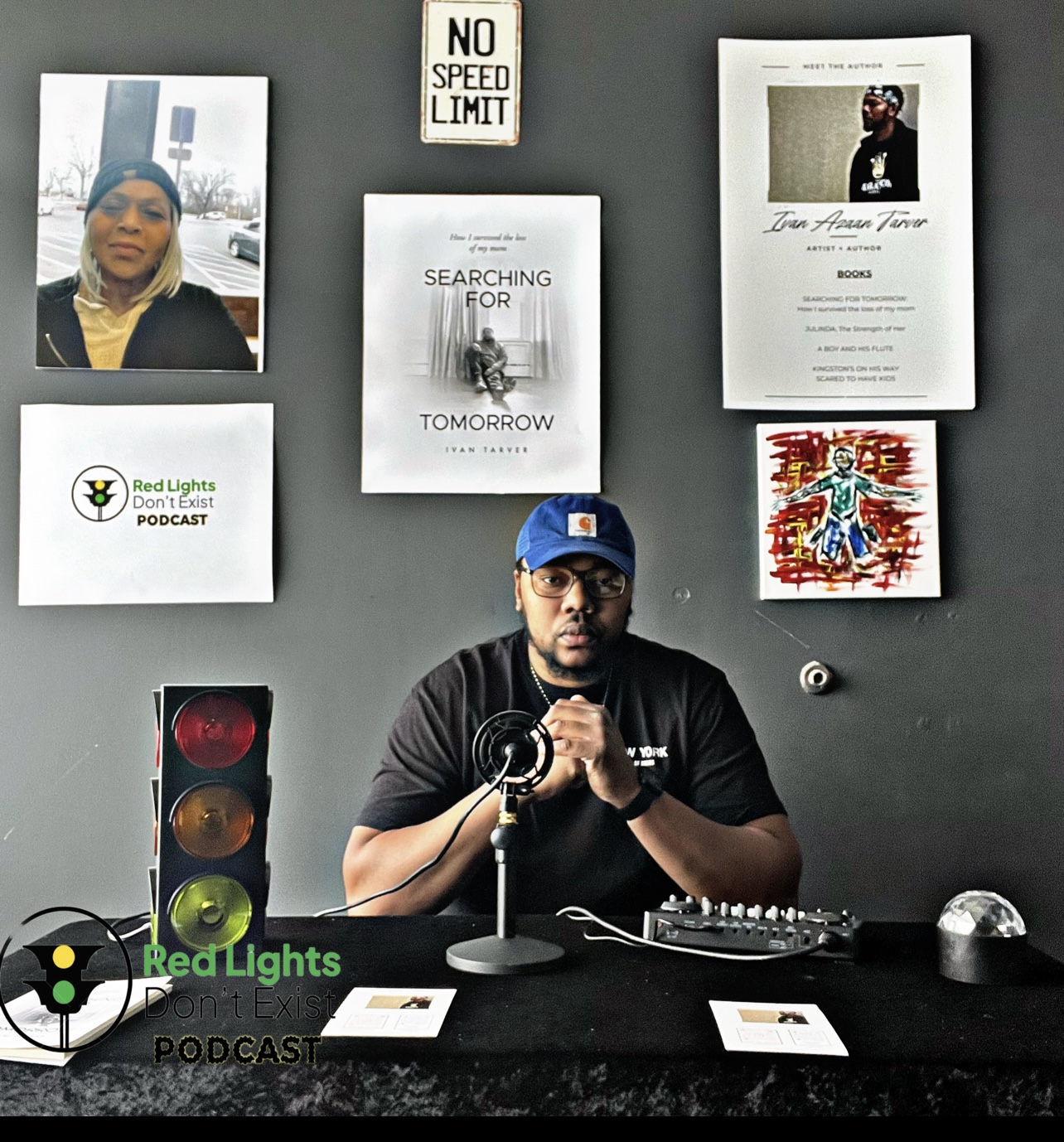 Man with a beard and glasses wearing a blue cap, sitting at a desk with podcast equipment, surrounded by posters and signs on a black wall.