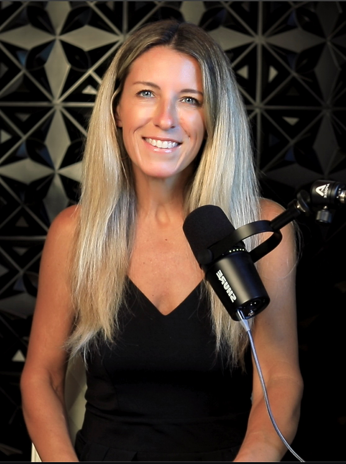 Woman with long blonde hair smiling, wearing a black top, in front of a geometric patterned background, speaking into a microphone.