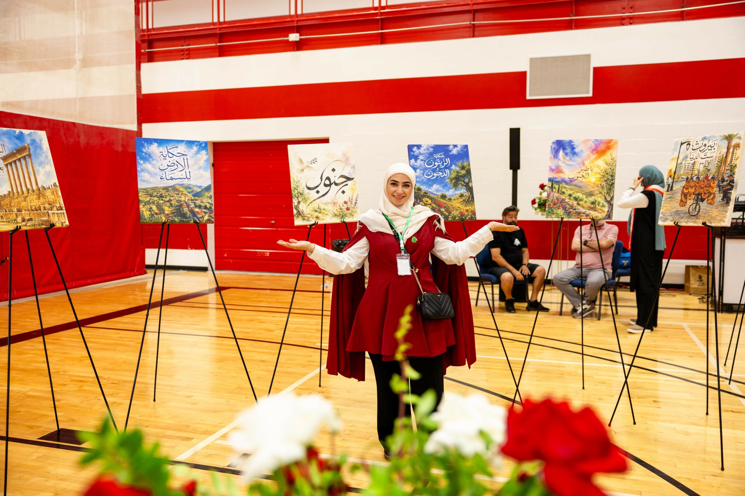 Woman in a white headscarf and maroon dress stands with arms outstretched in an art gallery with paintings on easels, wooden floor, red and white walls.