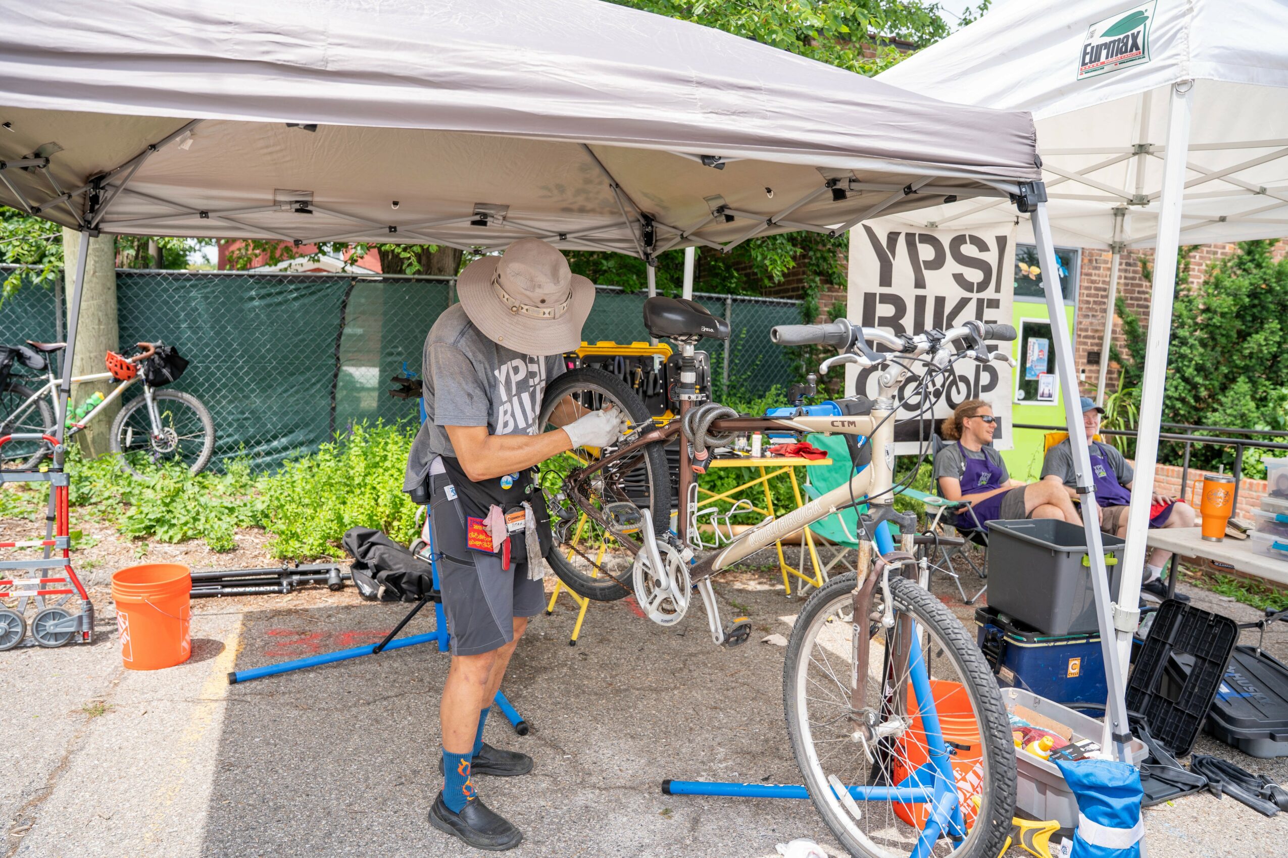 Person holding a bicycle under a canopy, with children and bicycles nearby, outdoor setting with trees and a fence.