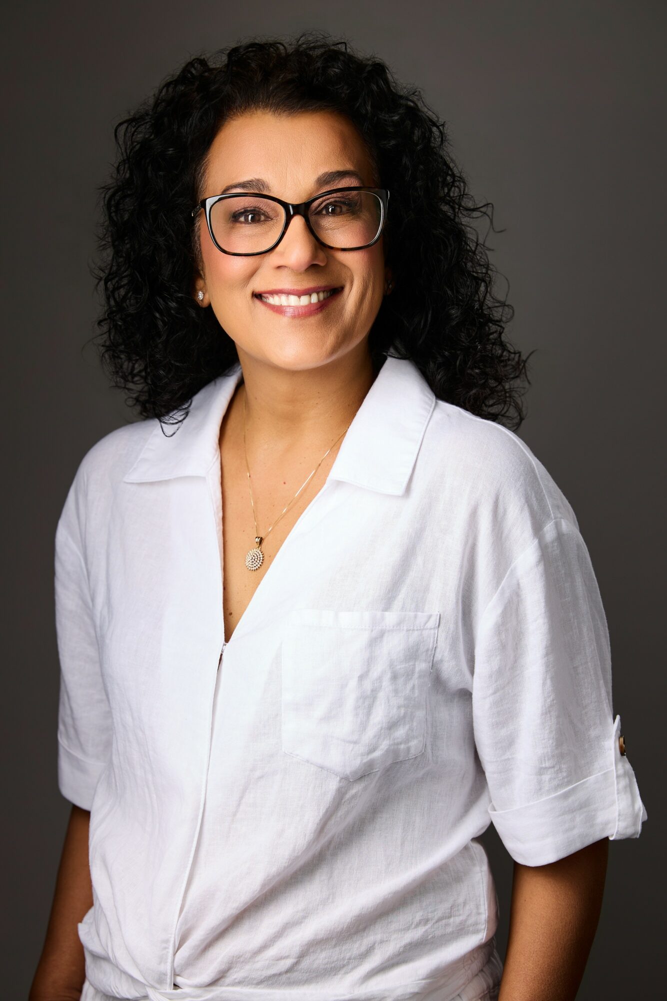 Woman with curly hair, glasses, smiling, wearing a white shirt against a dark background.