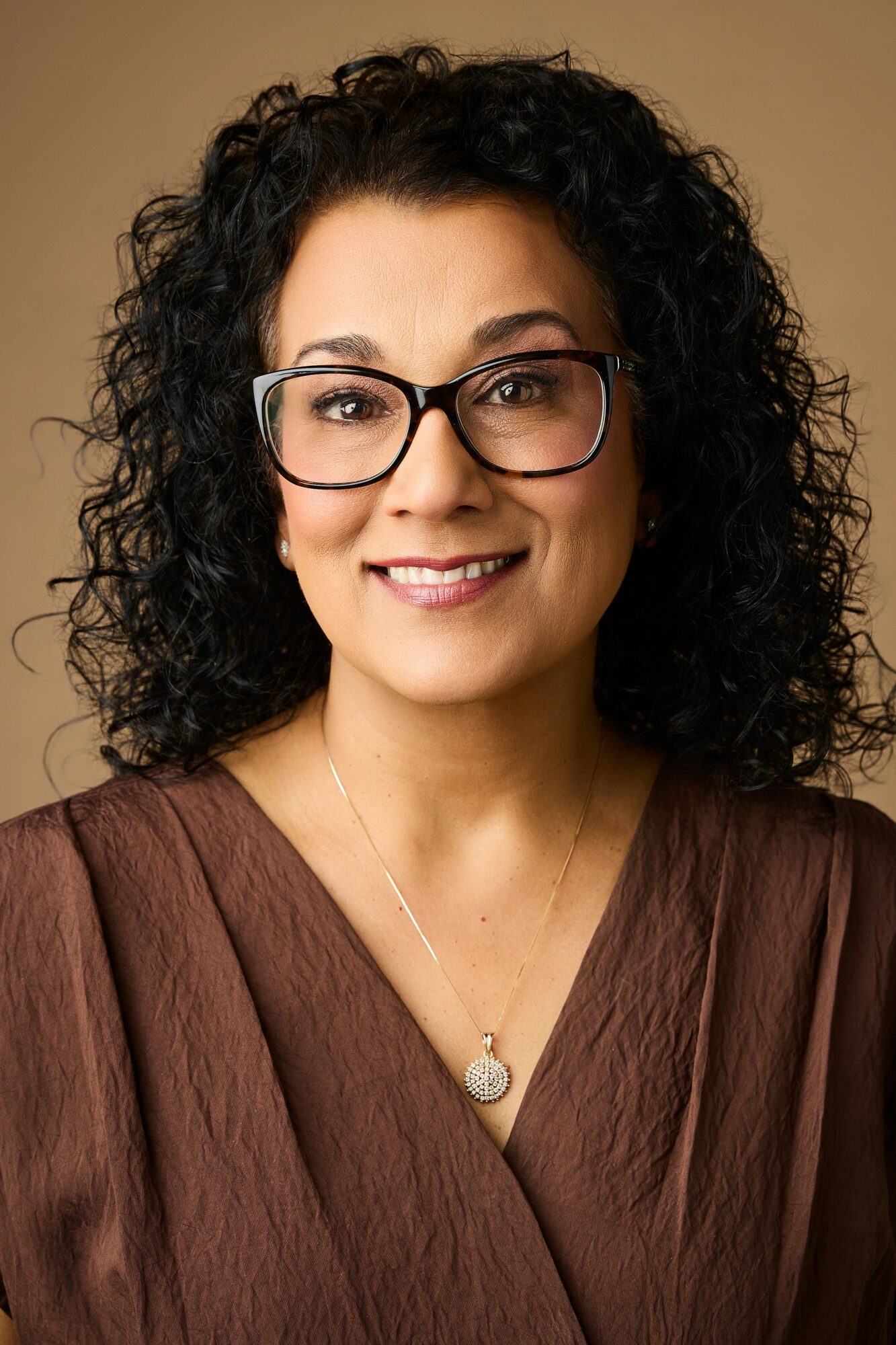 Woman with curly dark hair, glasses, smiling, wearing a brown top and necklace, against a neutral background.