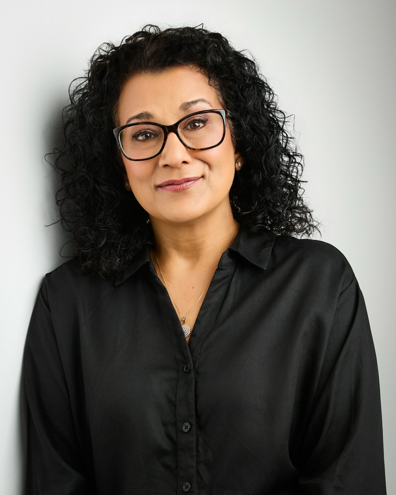 Woman with curly hair and glasses wearing a black shirt, smiling softly, standing against a light background.