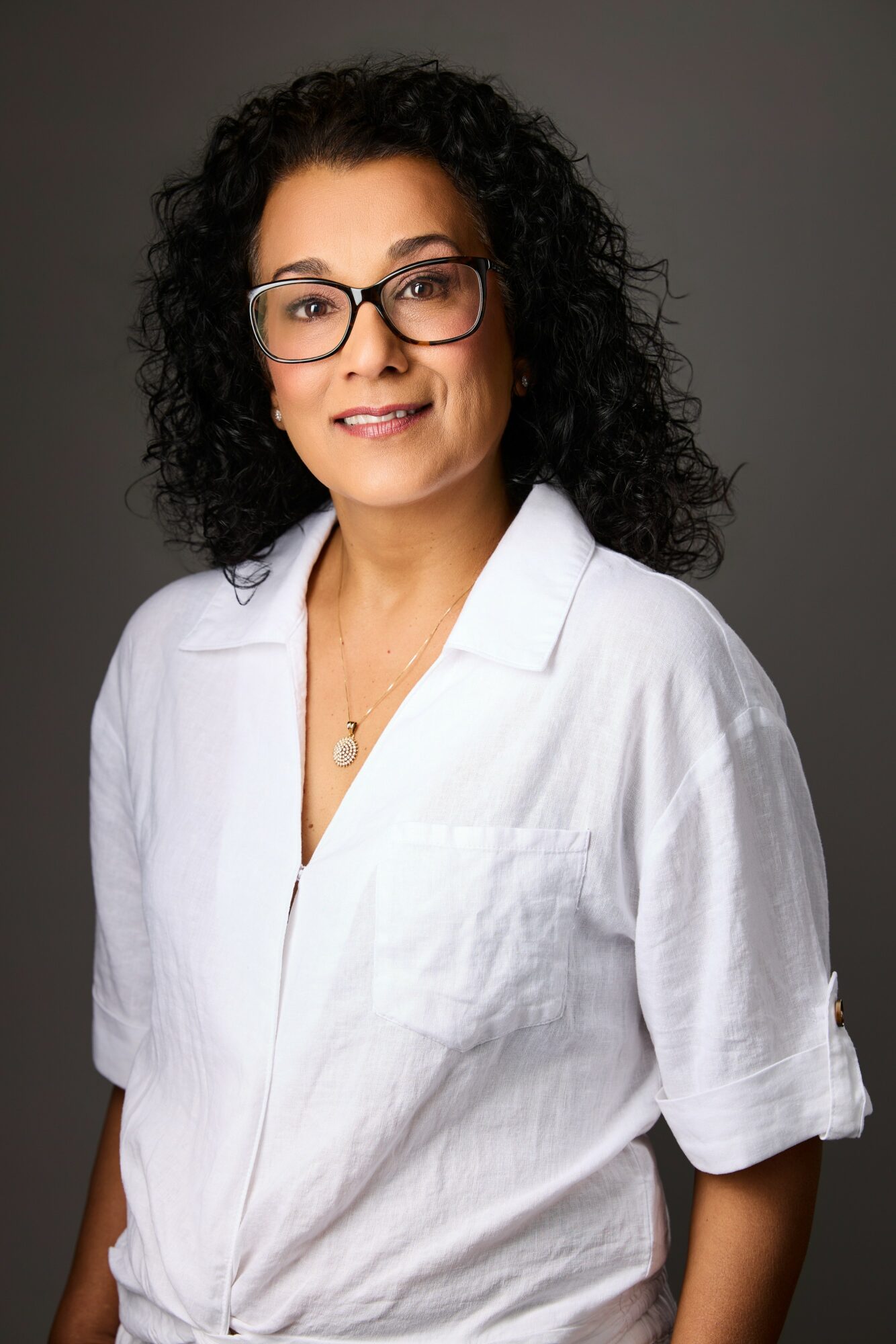 Woman with curly hair and glasses wearing a white shirt, smiling, against a gray background.