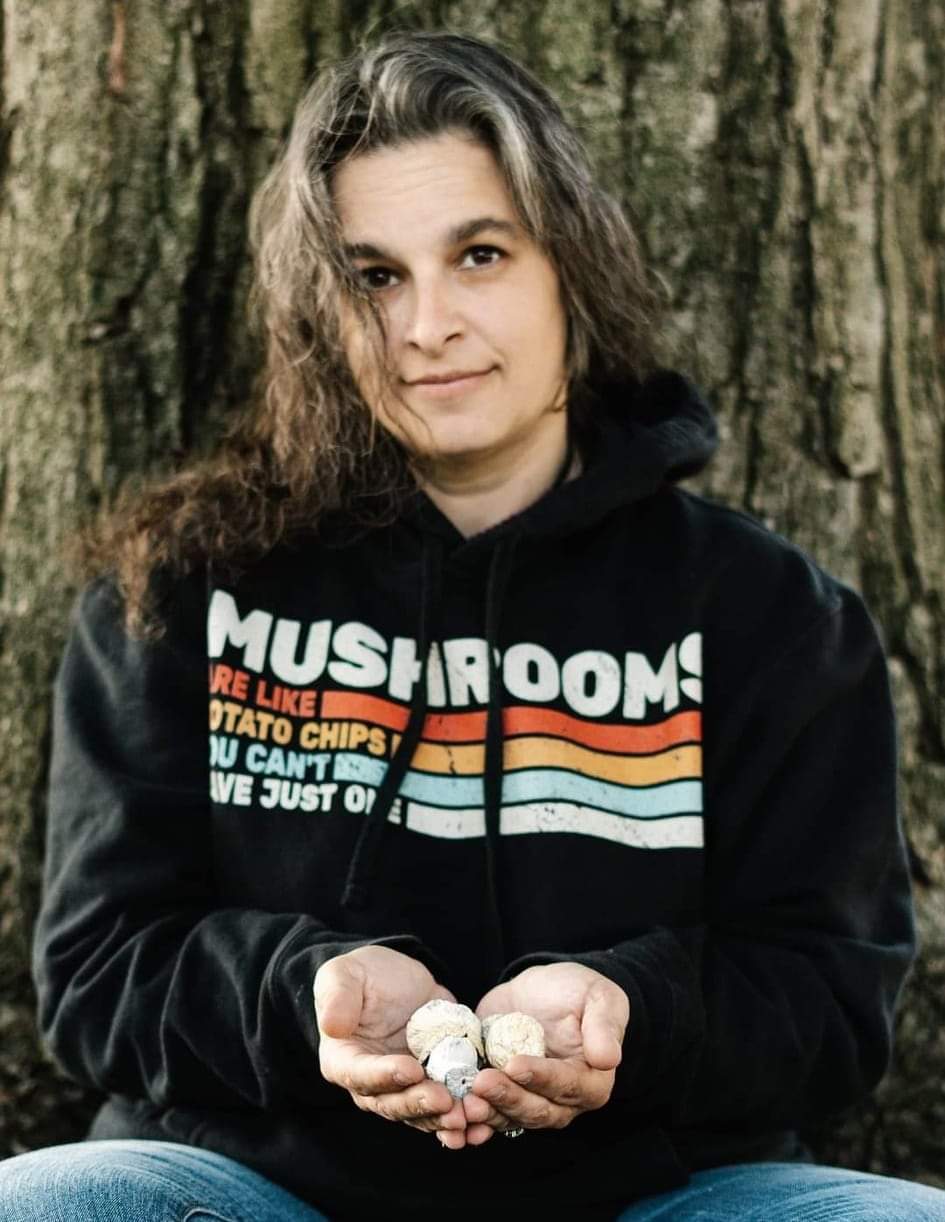 Young woman with wavy hair holding rocks in front of a tree, wearing a black hoodie with colorful stripes and text.