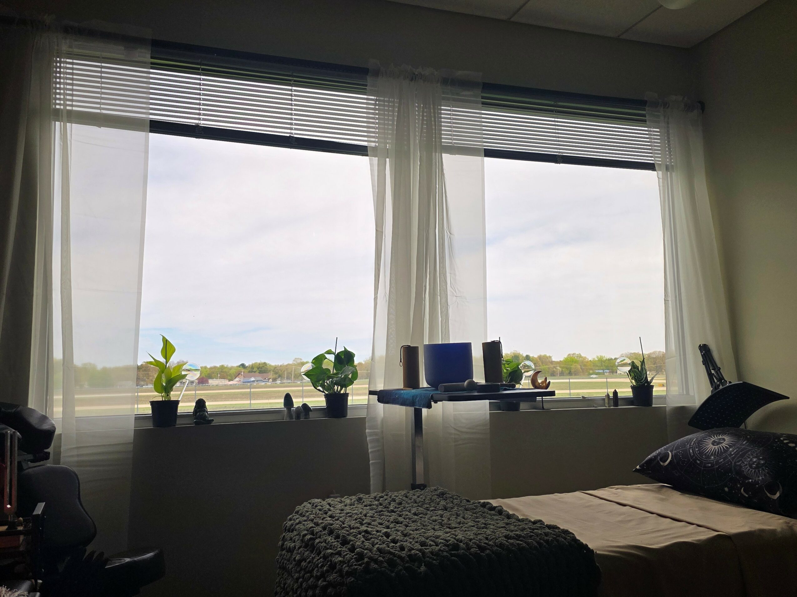 Room with large window, sheer curtains, plants on windowsill, bed with pillow, and an ironing board inside.