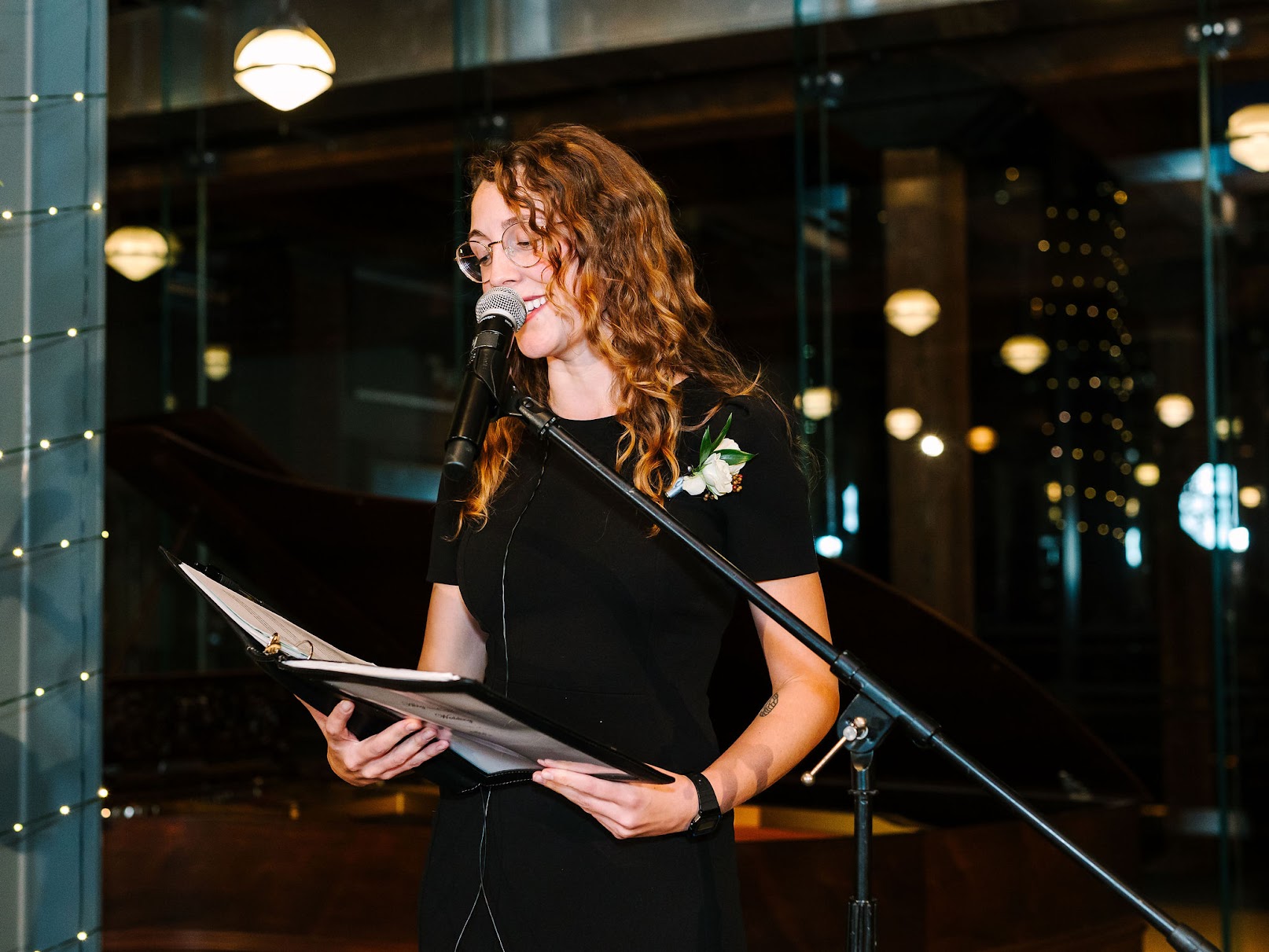 Woman with curly hair reading from a book at a microphone, indoors with reflections of lights in the background.