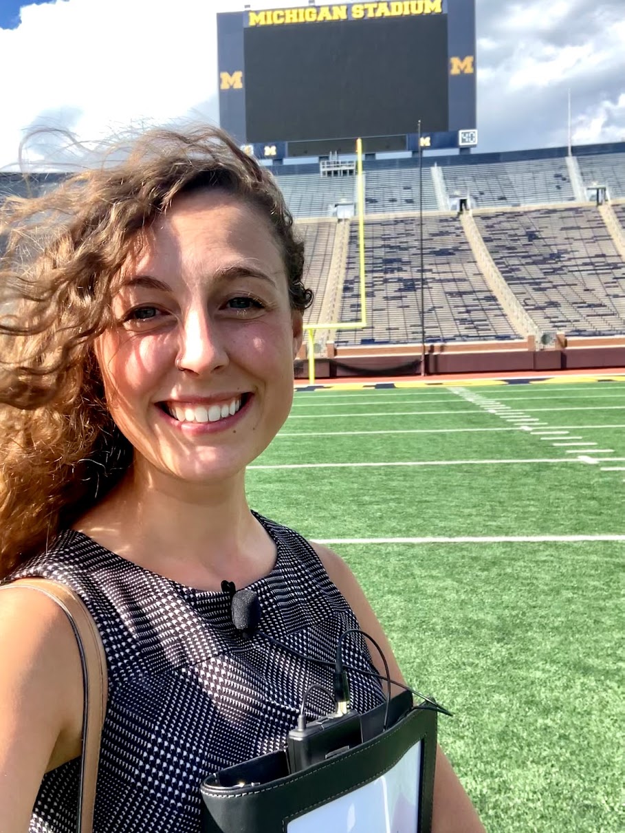 Smiling woman with curly hair taking selfie at Michigan Stadium, empty seats and scoreboard in background.