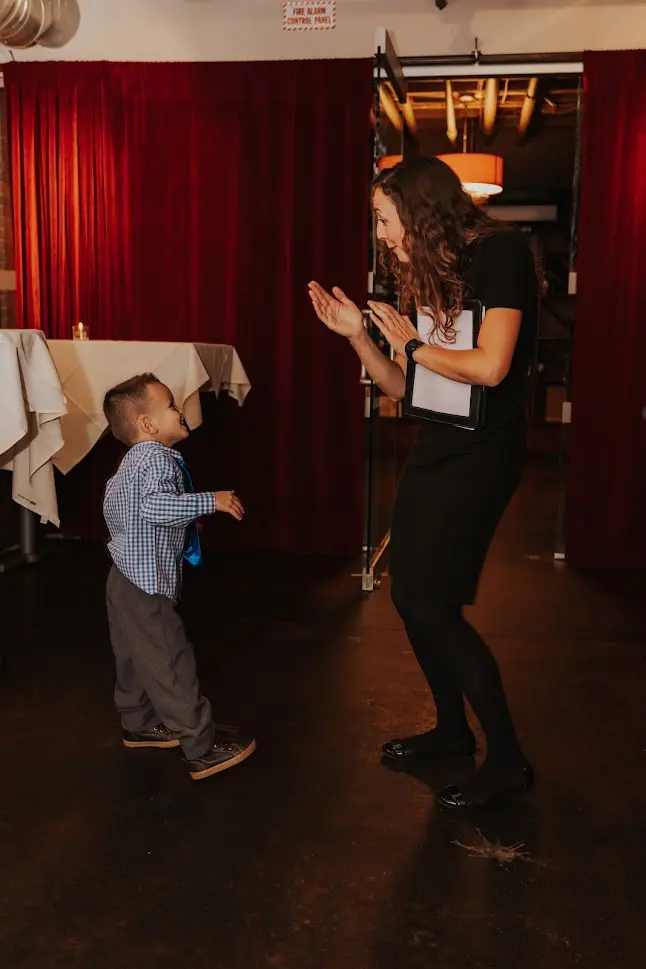 Young woman and small boy interacting indoors, woman clapping hands, boy looking up, dimly lit room with red curtains and tables.