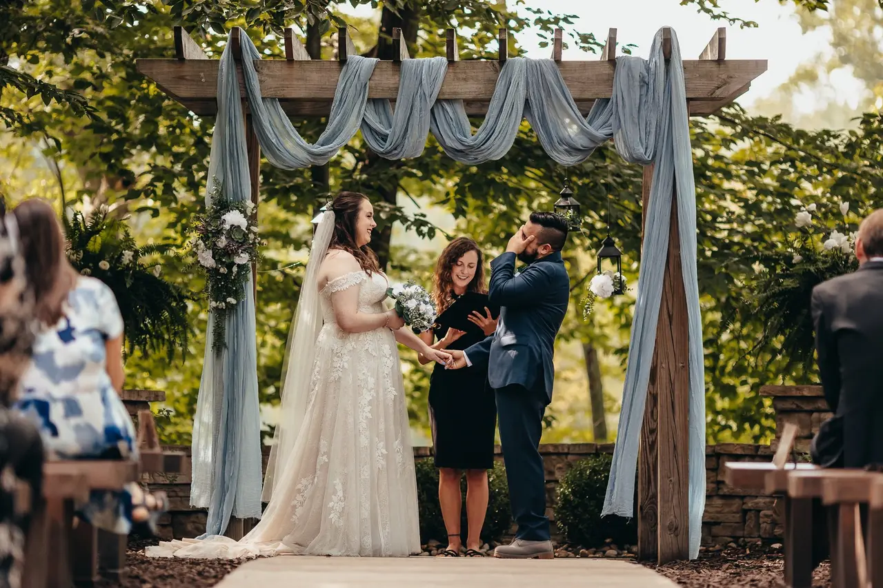 Bride and groom exchanging vows under a decorated wooden arch with an officiant, outdoors with trees and sunlight, guests seated nearby.
