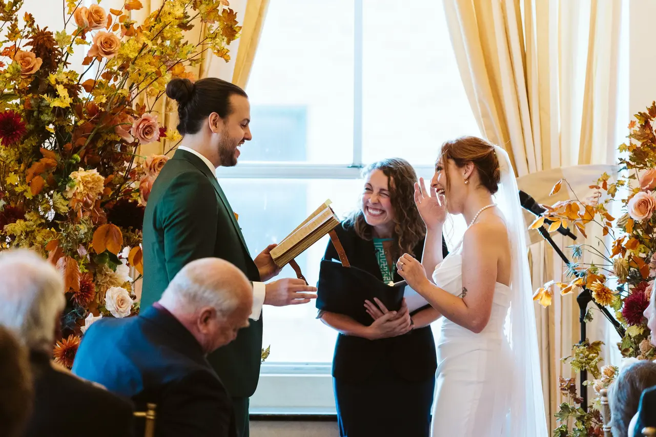 Man in suit and woman in wedding dress exchange vows with a woman officiant, surrounded by flowers and guests.