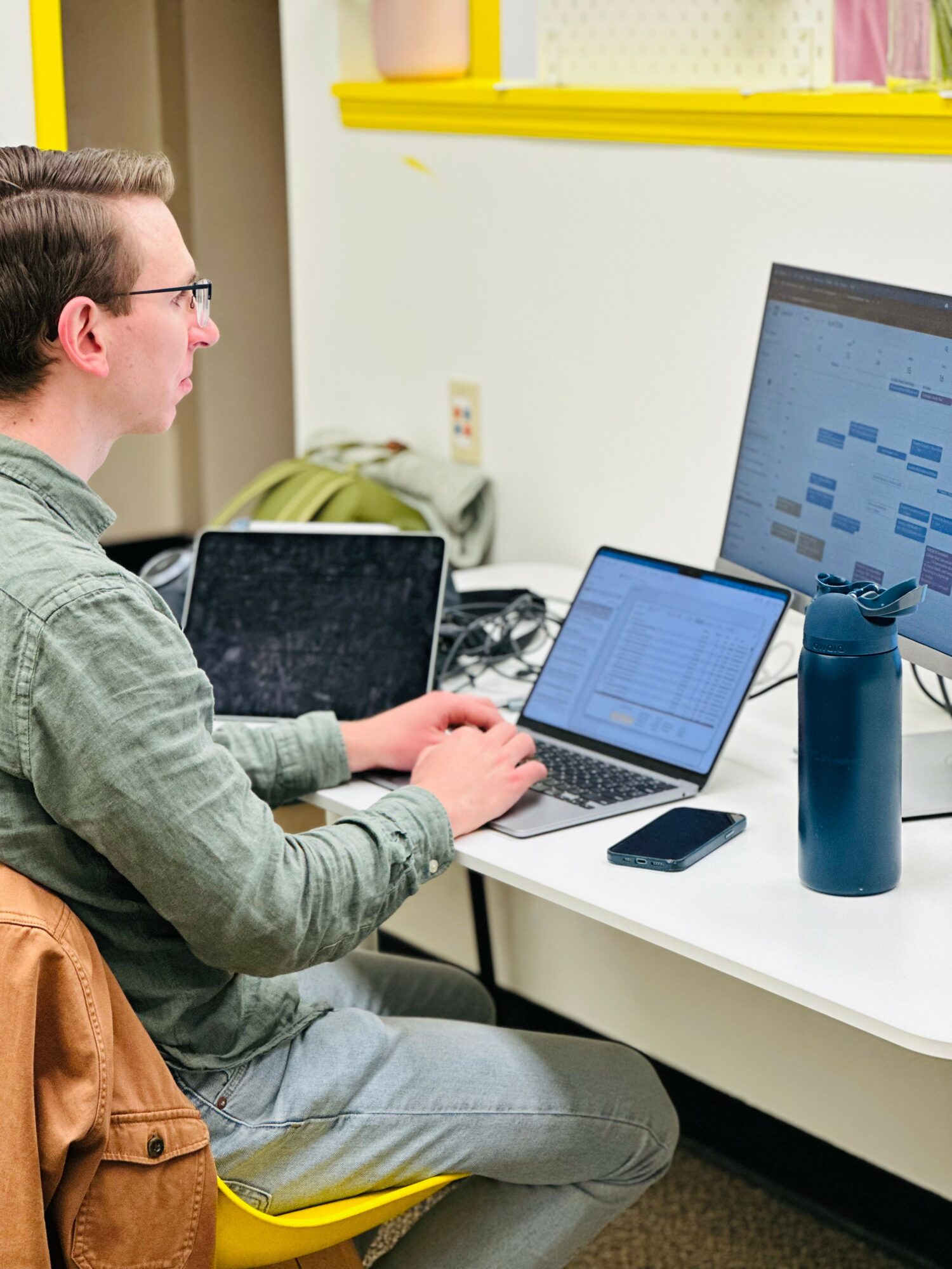 Person working on multiple laptops at a desk with a large monitor, water bottle, and phone, in a room with yellow trim.