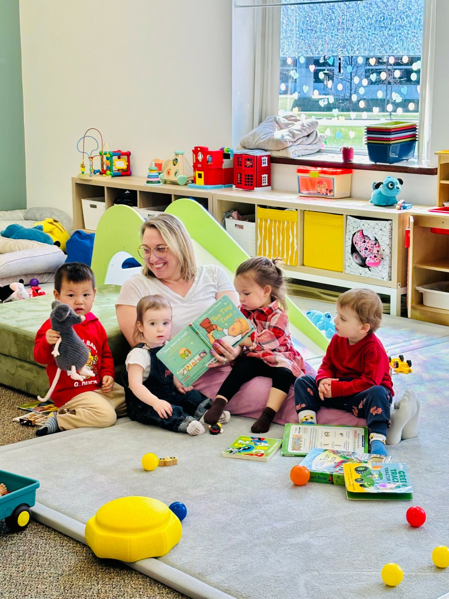 Adult woman with four children sitting on a play mat in a colorful playroom, surrounded by toys and books, near a window.