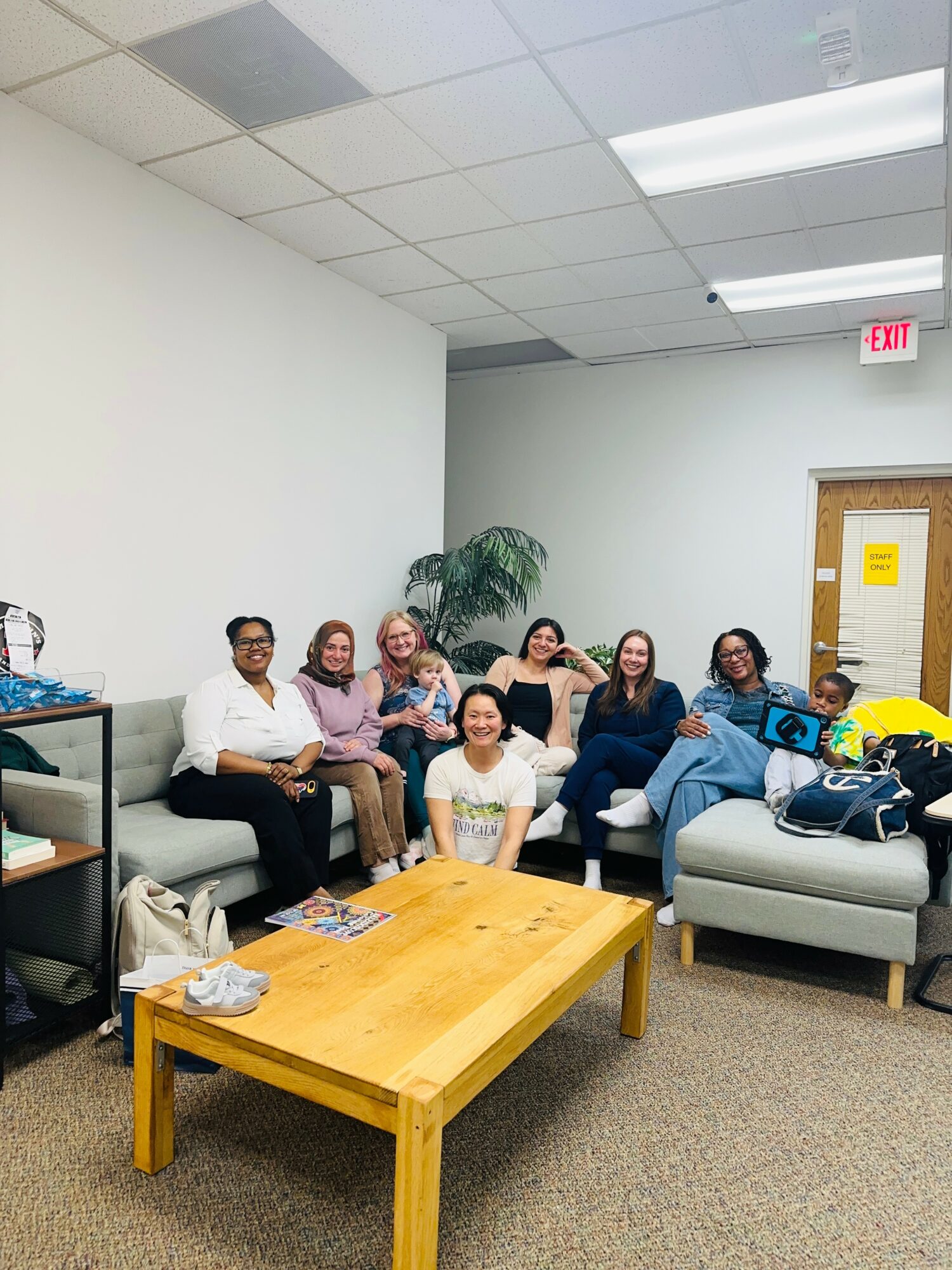 Group of seven women sitting on couches in a room with white walls and ceiling, a wooden table in front, and a door in the background.