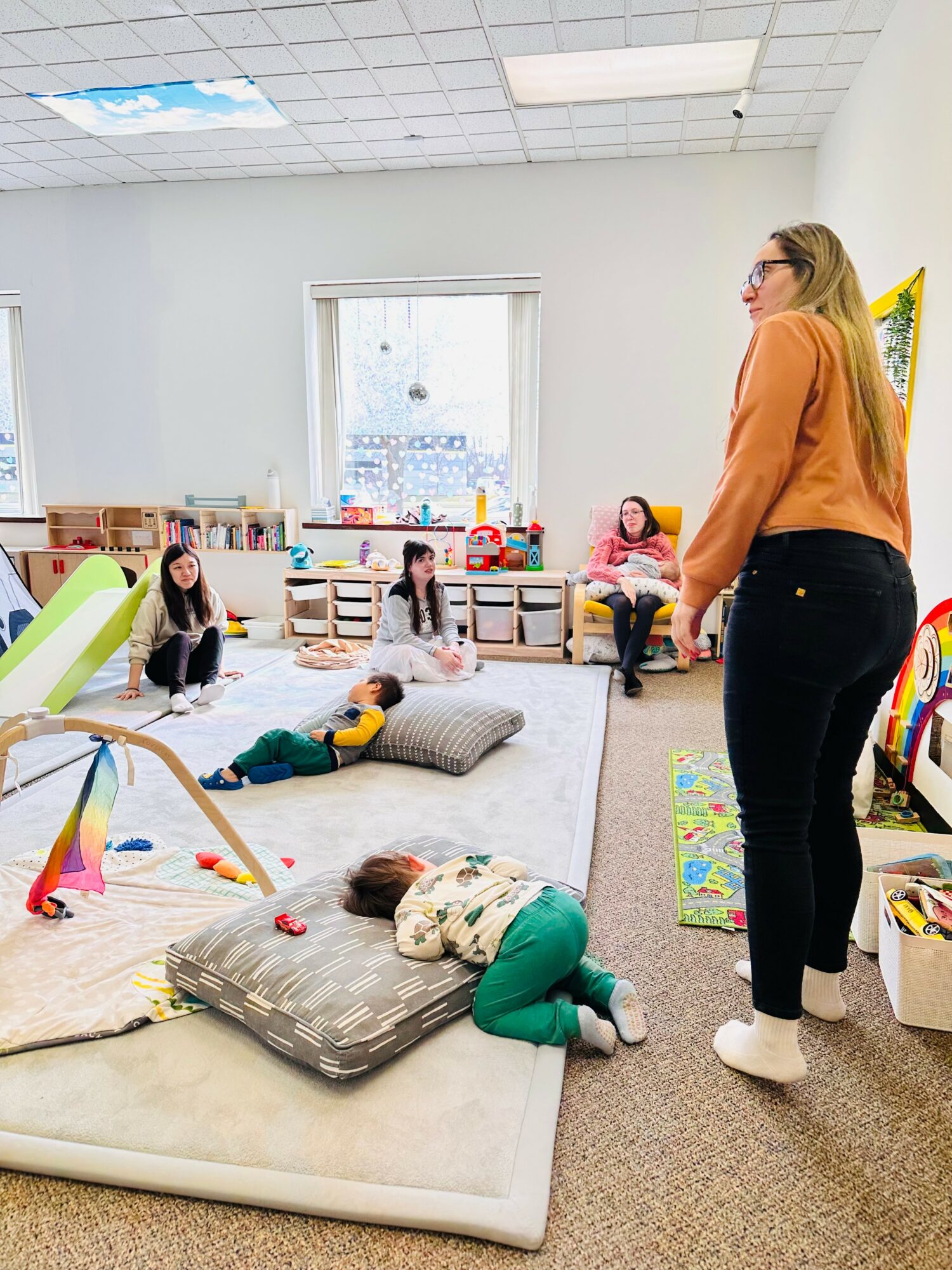 Classroom with children and teacher, some children lying on mats, teacher standing, colorful toys and books, window with blinds.