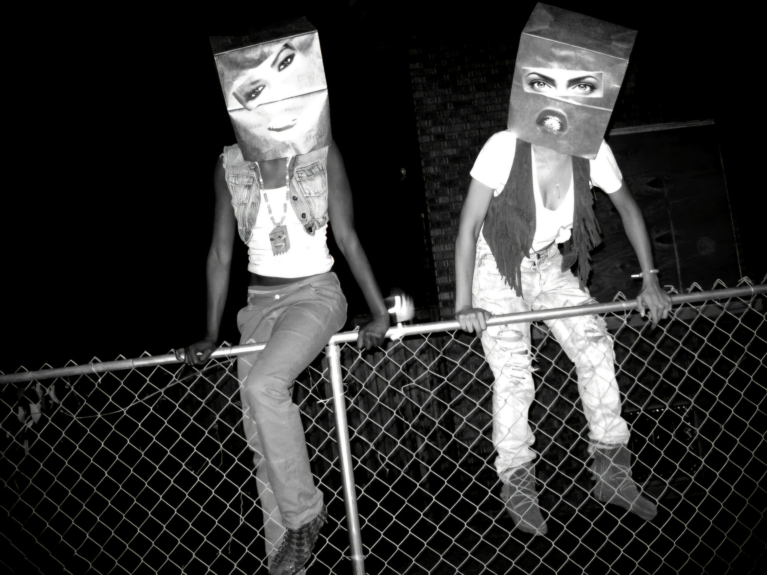 Two people wear paper bags with eye and mouth cutouts, standing on a chain-link fence at night.