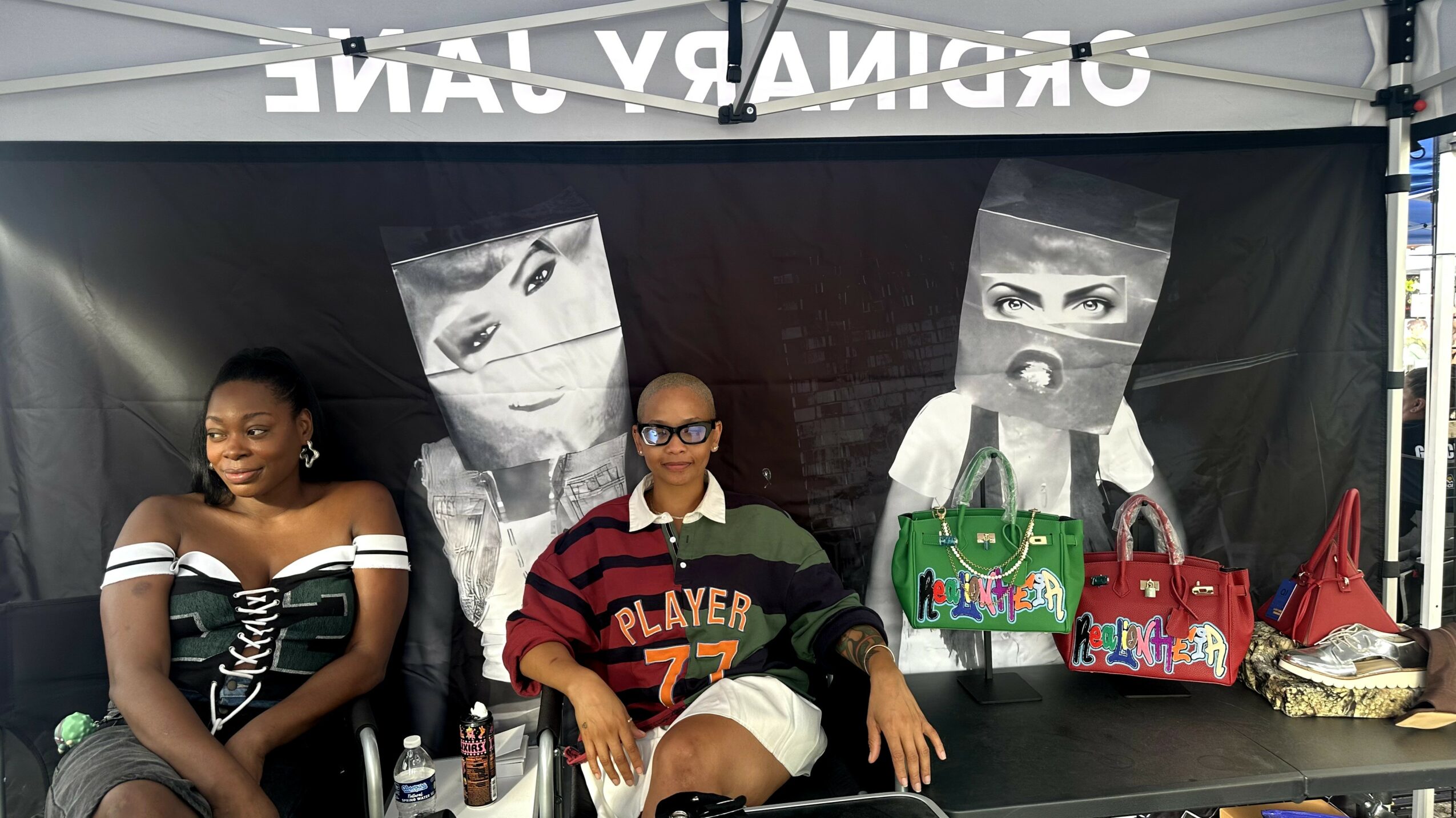 Three women sitting at a table with bags and a backdrop featuring black-and-white faces.