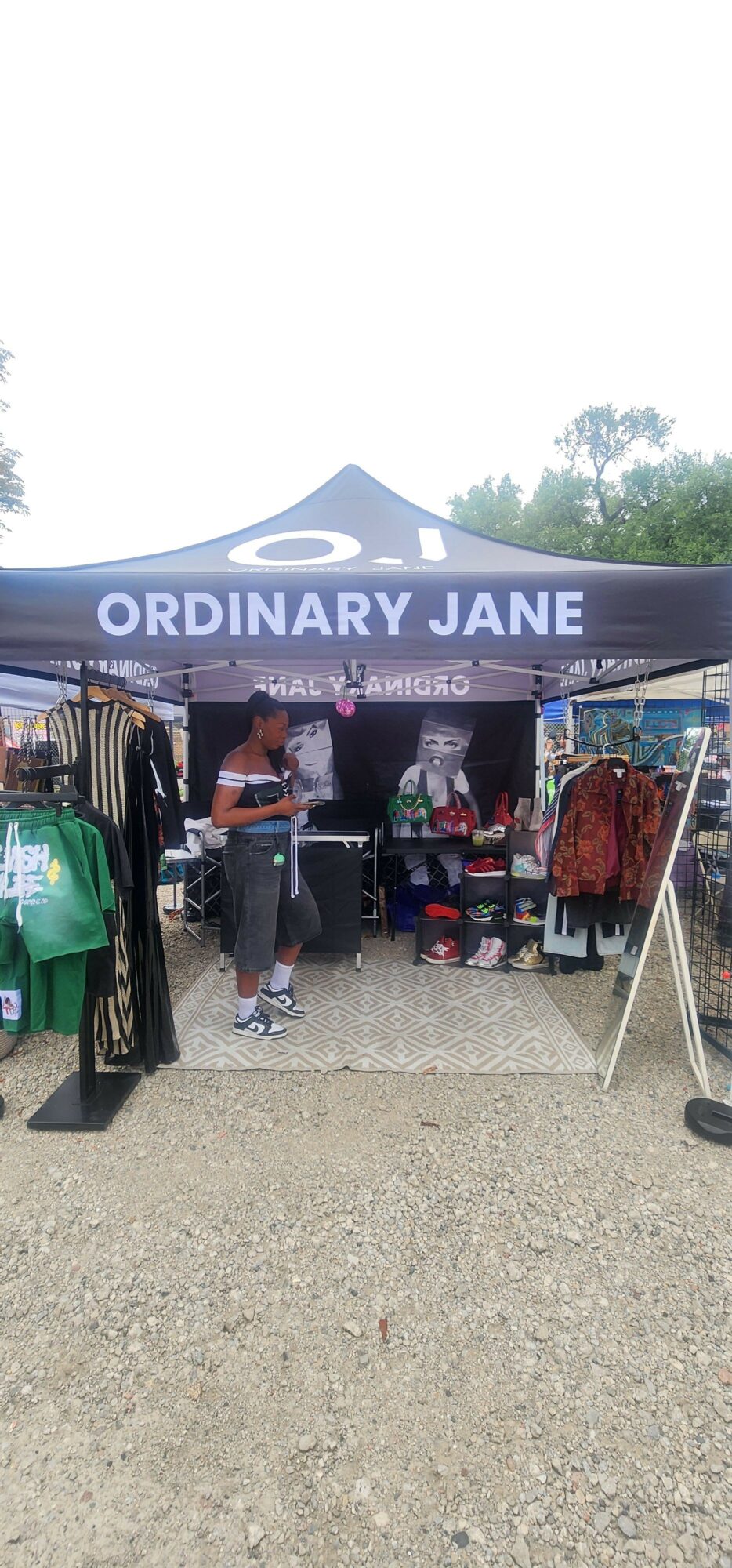 People under a tent with a sign reading 'ORDINARY JANE' at an outdoor event, with bags and items nearby.