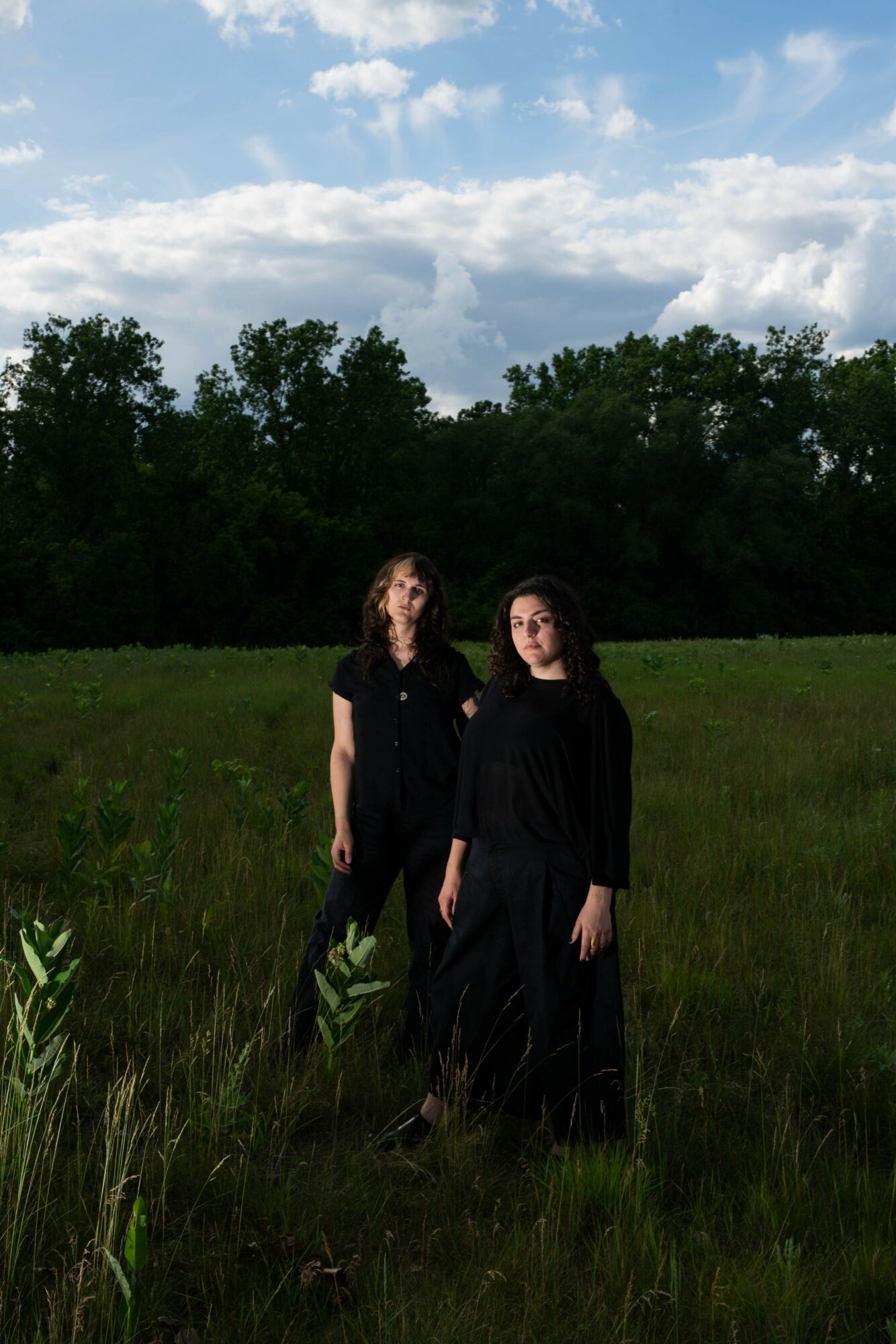 Two women standing in a grassy field with trees and a partly cloudy sky in the background.