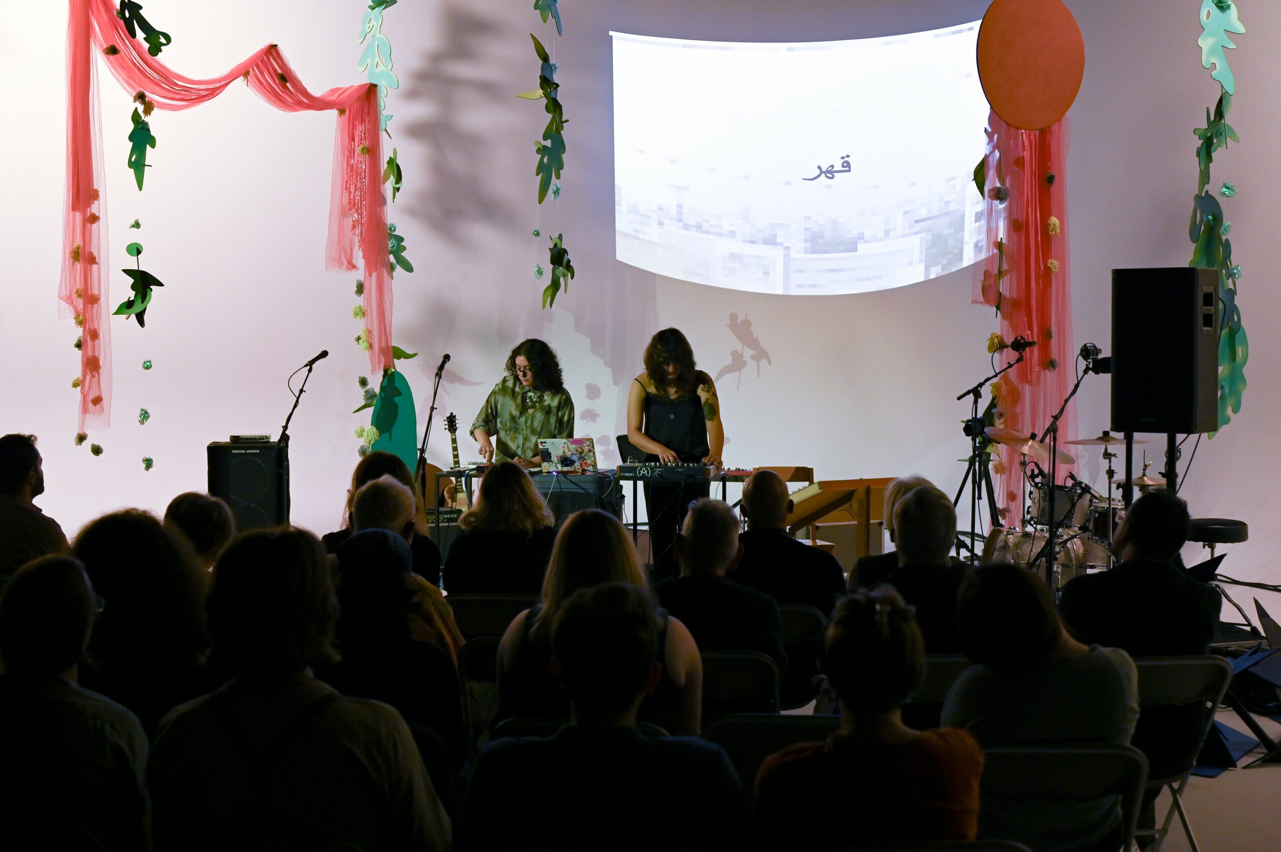 Two performers on stage with musical instruments, audience watching, decorated with pink and green streamers, large screen behind.