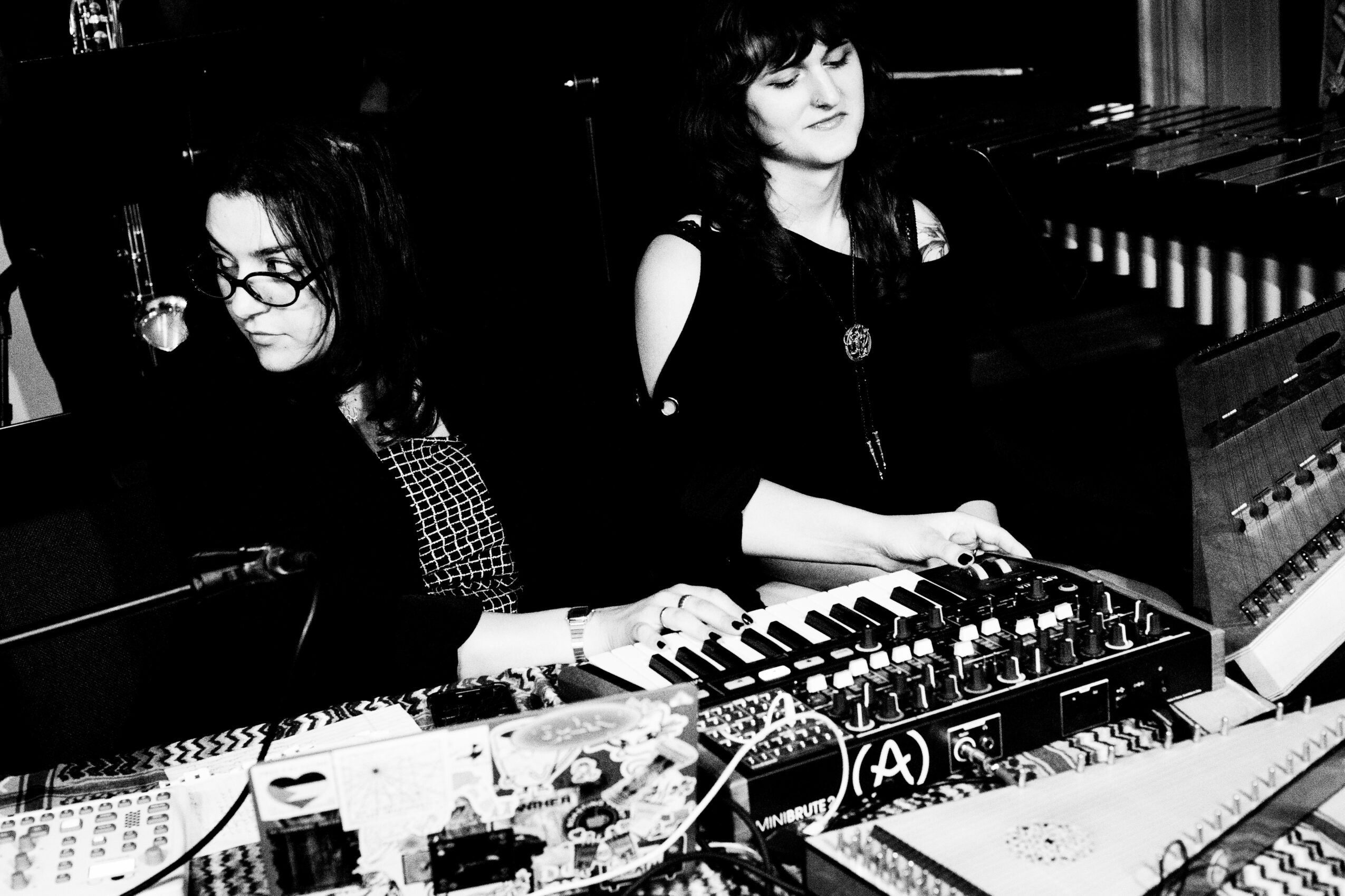 Two women are playing keyboard instruments in a dark room, one with glasses and the other with dark hair, surrounded by music equipment.