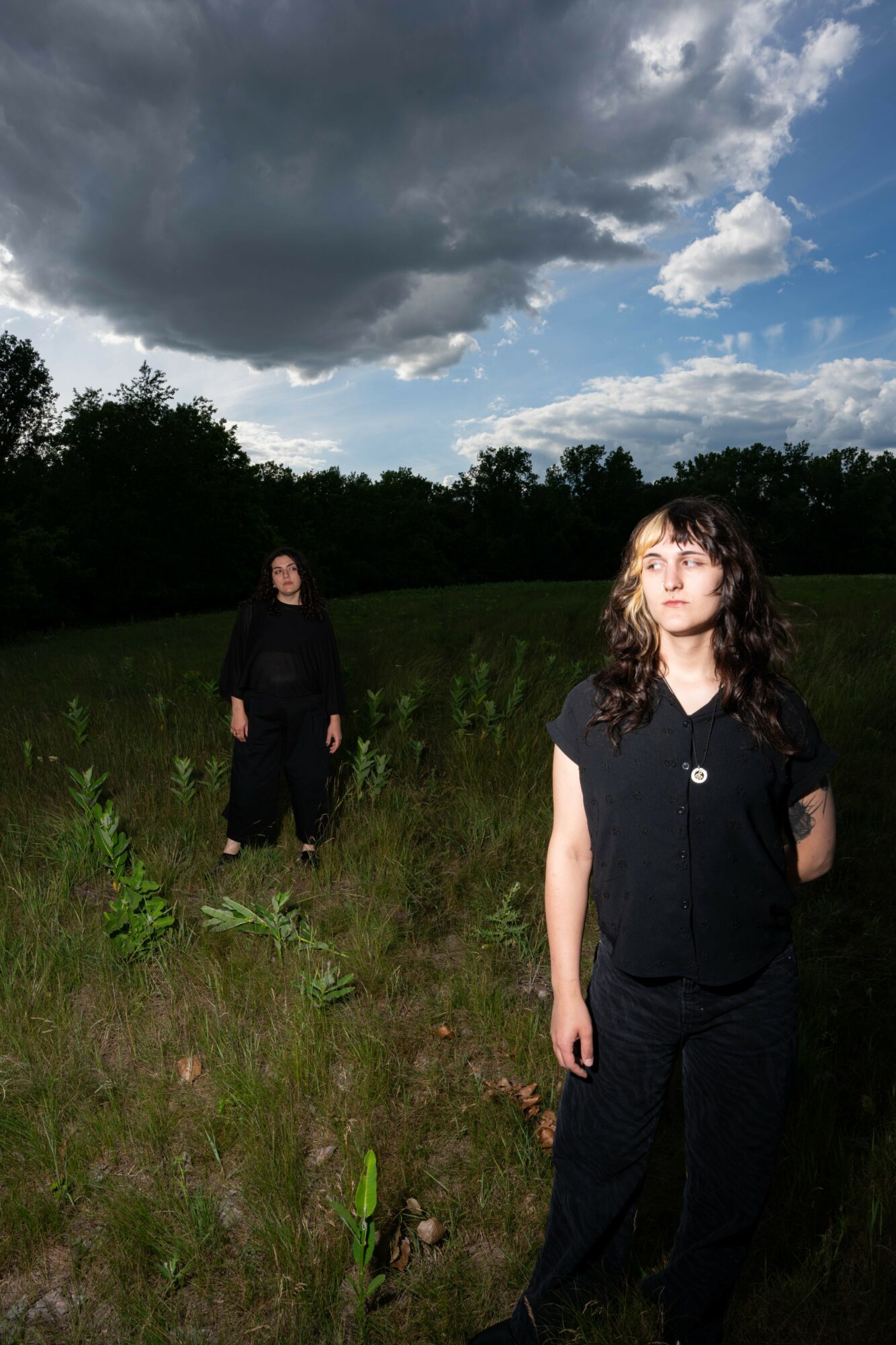 Two women standing in a grassy field under a cloudy sky, one in the foreground and one in the background.