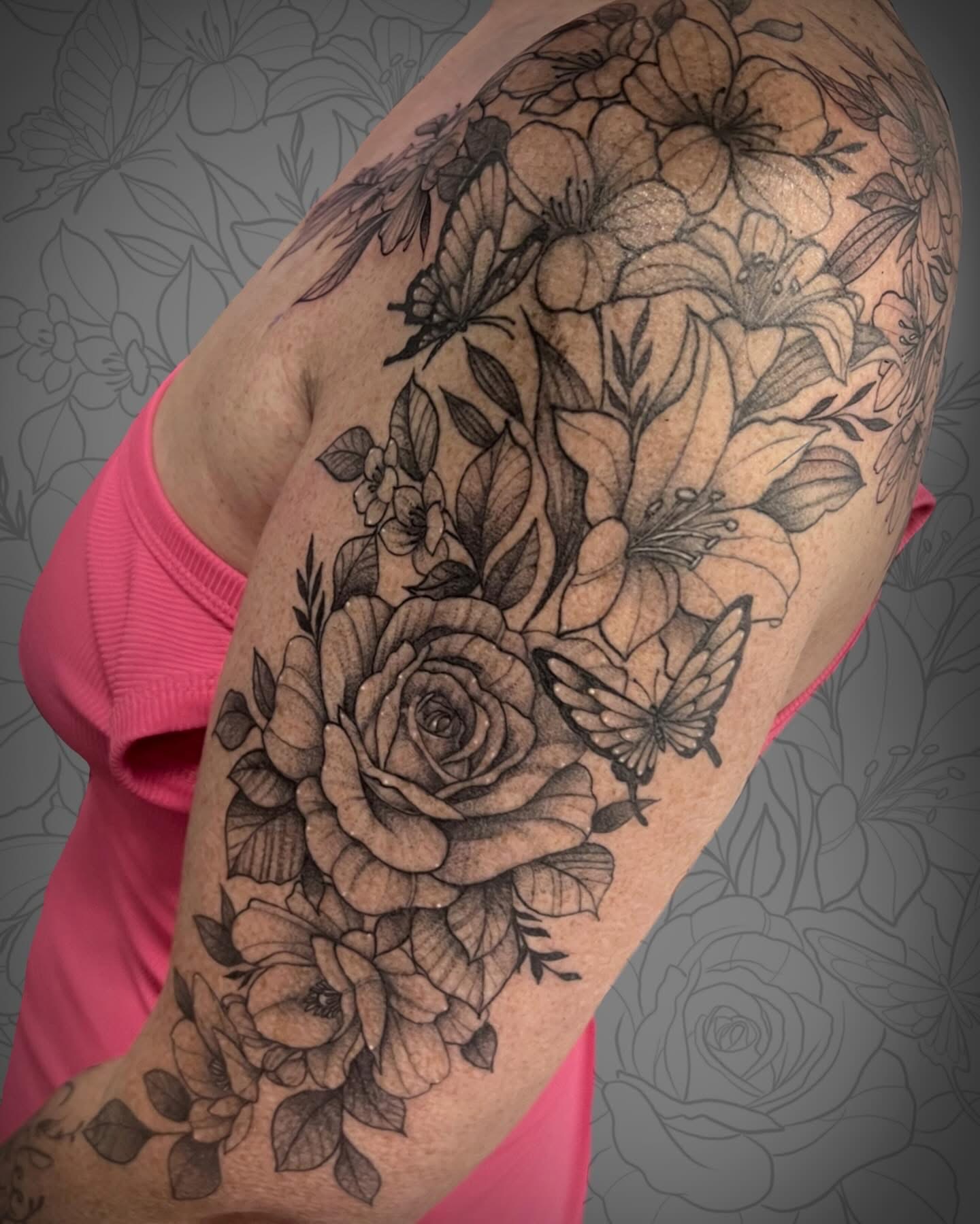 Tattoo of flowers, a butterfly, and a rose on person's upper arm, with pink clothing visible, background with floral pattern.
