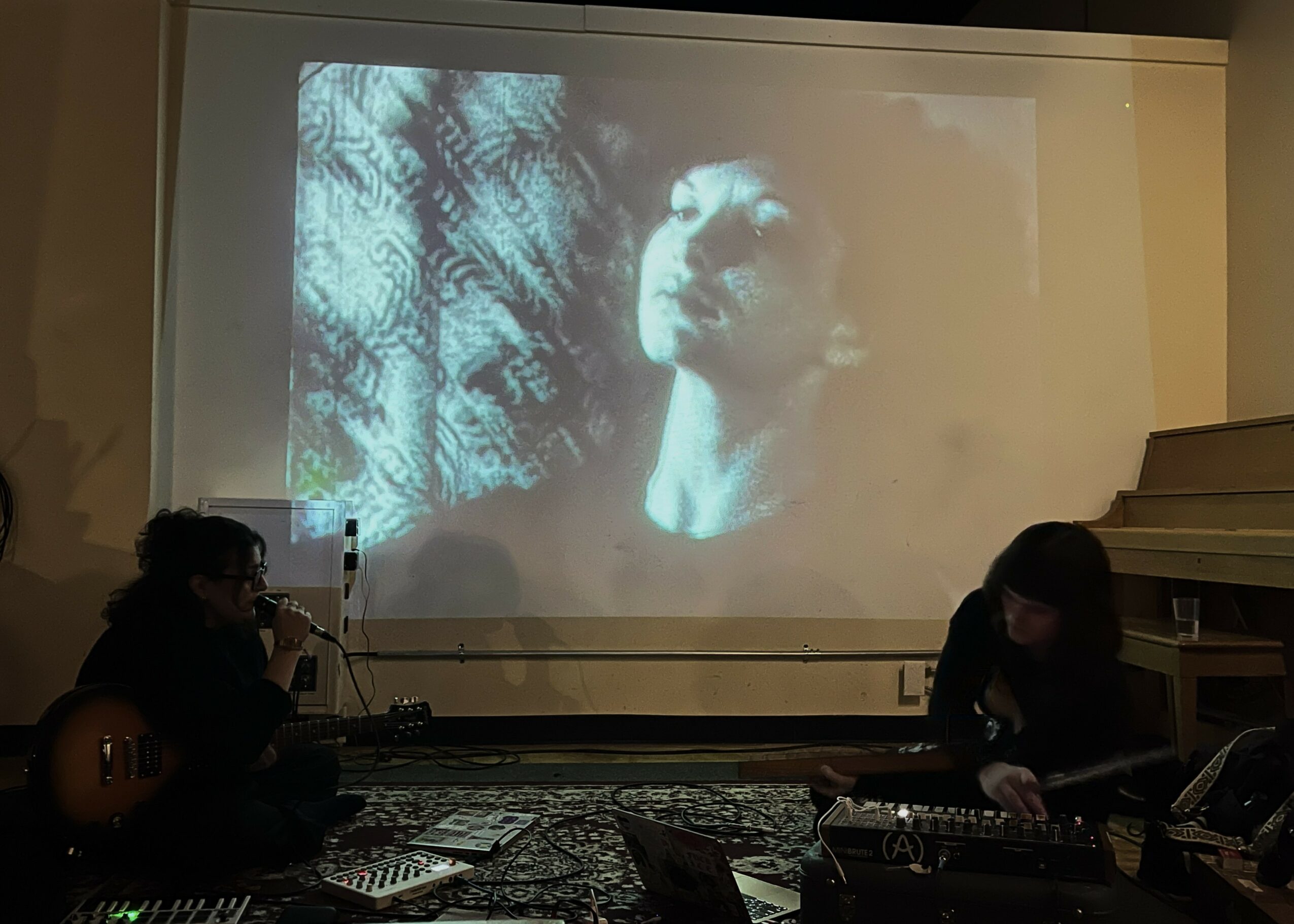 Two people perform music in front of a projected black-and-white film of a person's face, with a patterned background.