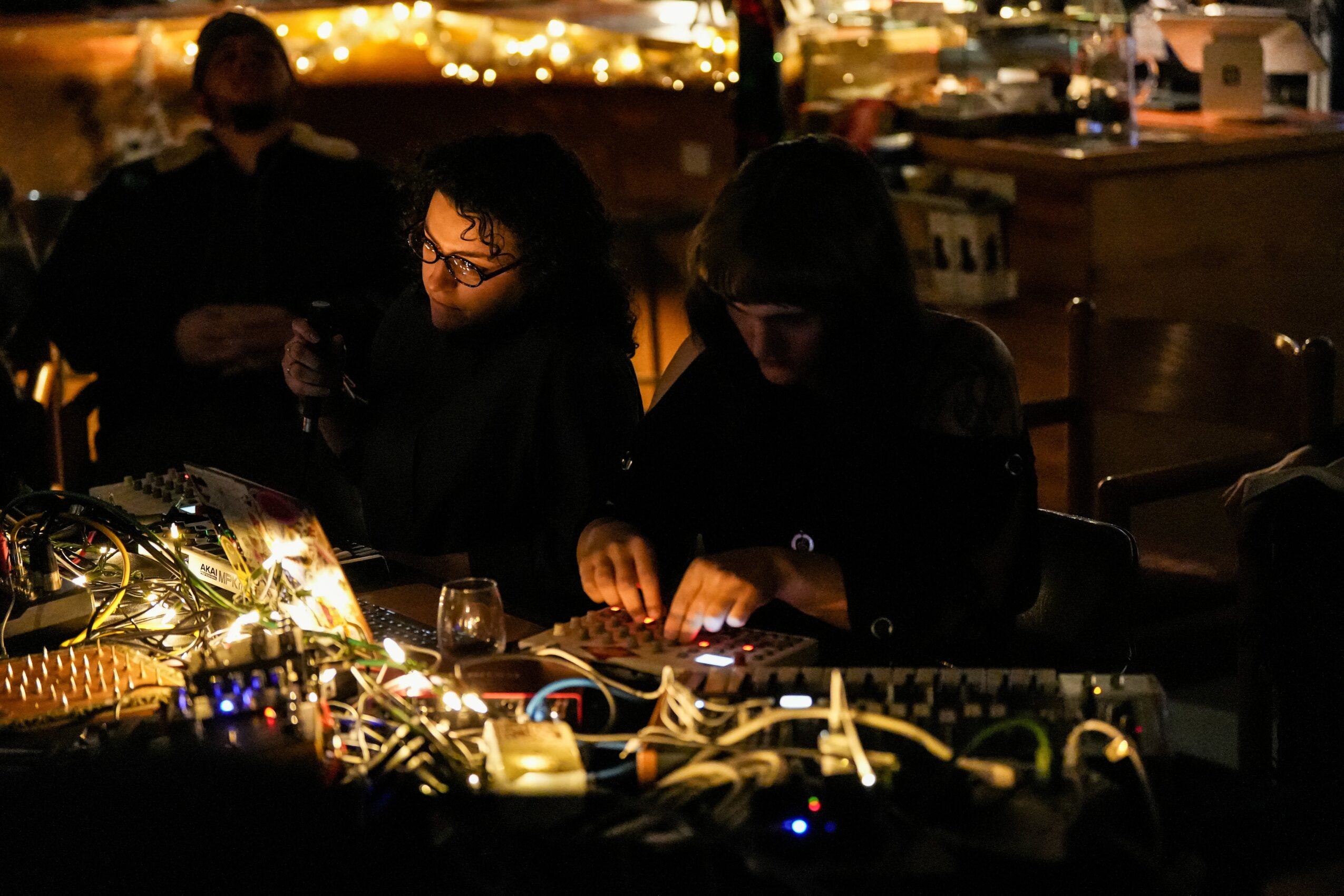 Two women sit at a table with electronic equipment, illuminated by string lights, in a nighttime outdoor setting.