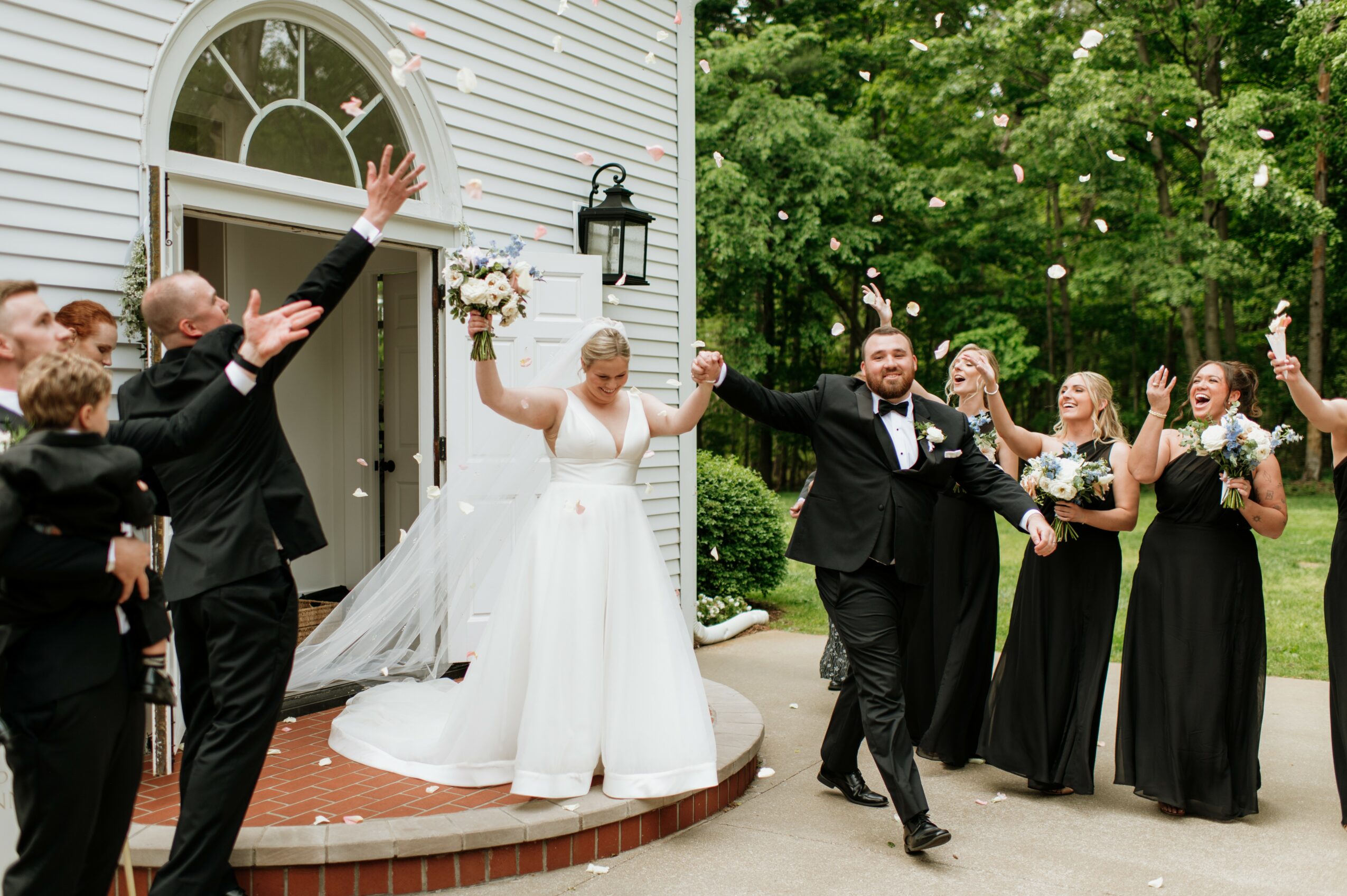 Bride and groom celebrating outside with friends, some raising hands, in front of a white building with greenery.