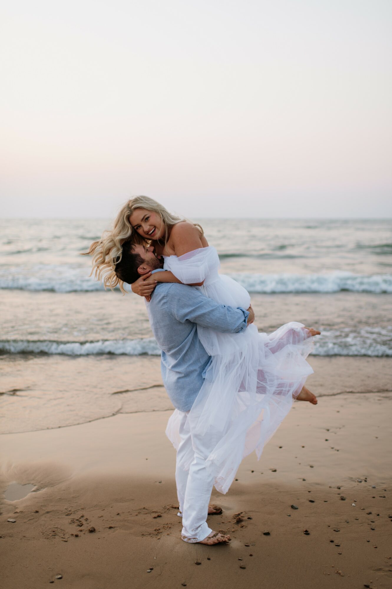 A couple on the beach, the woman in a white dress being carried by the man in a light blue shirt, ocean in background