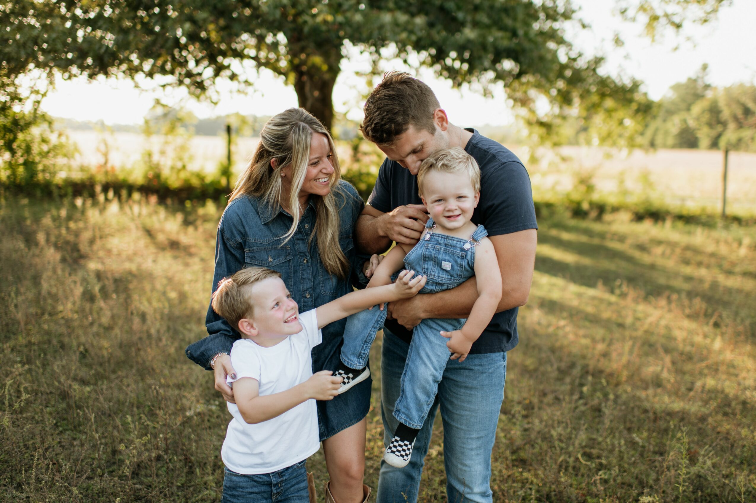 Family of four outdoors, smiling and playing together in a grassy area with trees in background.