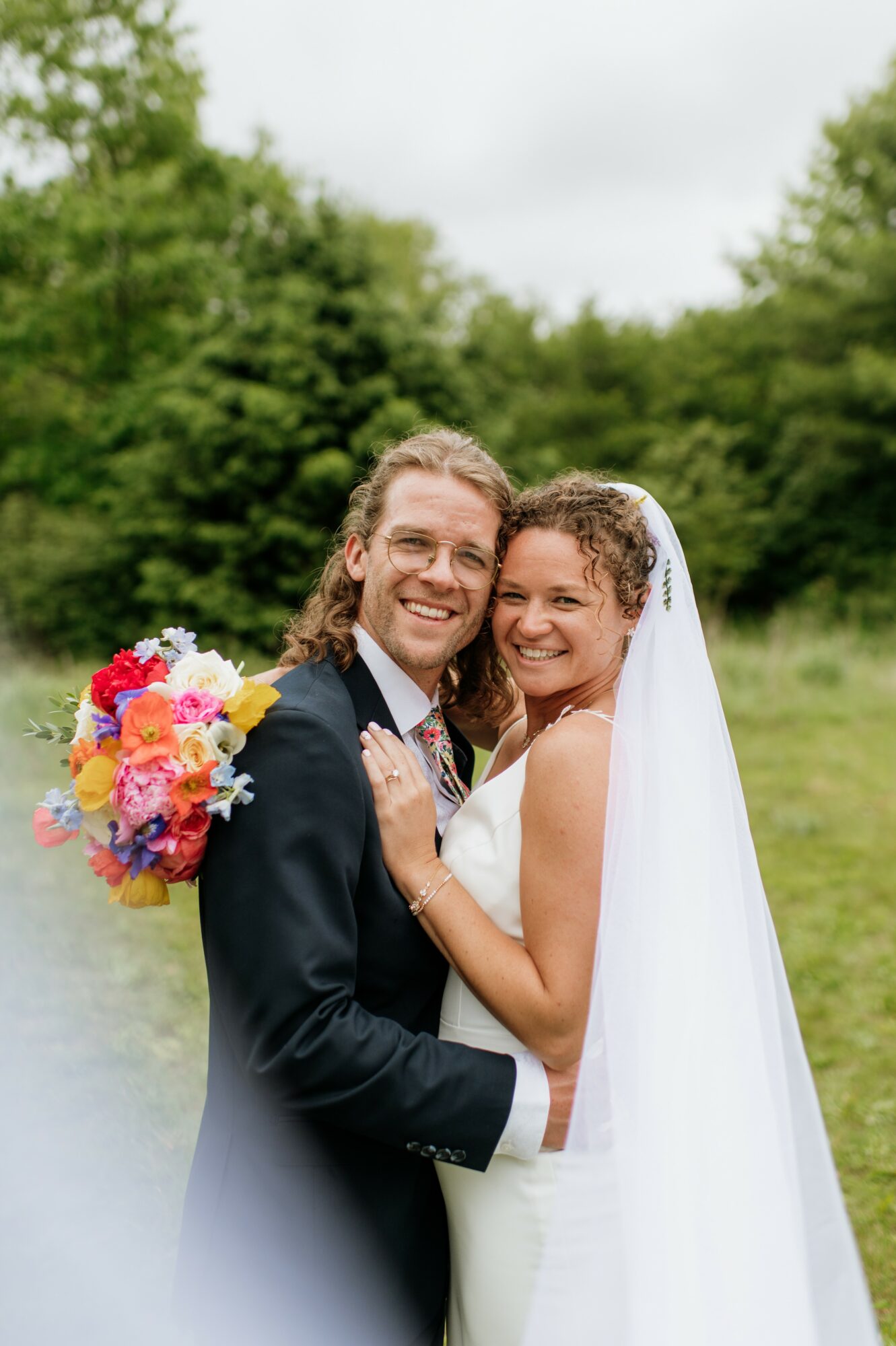 A smiling bride and groom embrace outdoors, with trees and cloudy sky in the background.