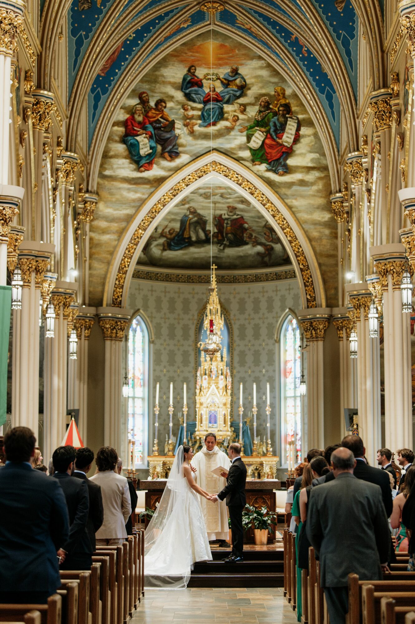 Wedding ceremony inside a grand church with high vaulted ceiling, ornate gold details, and stained glass windows, couple at altar, guests seated.