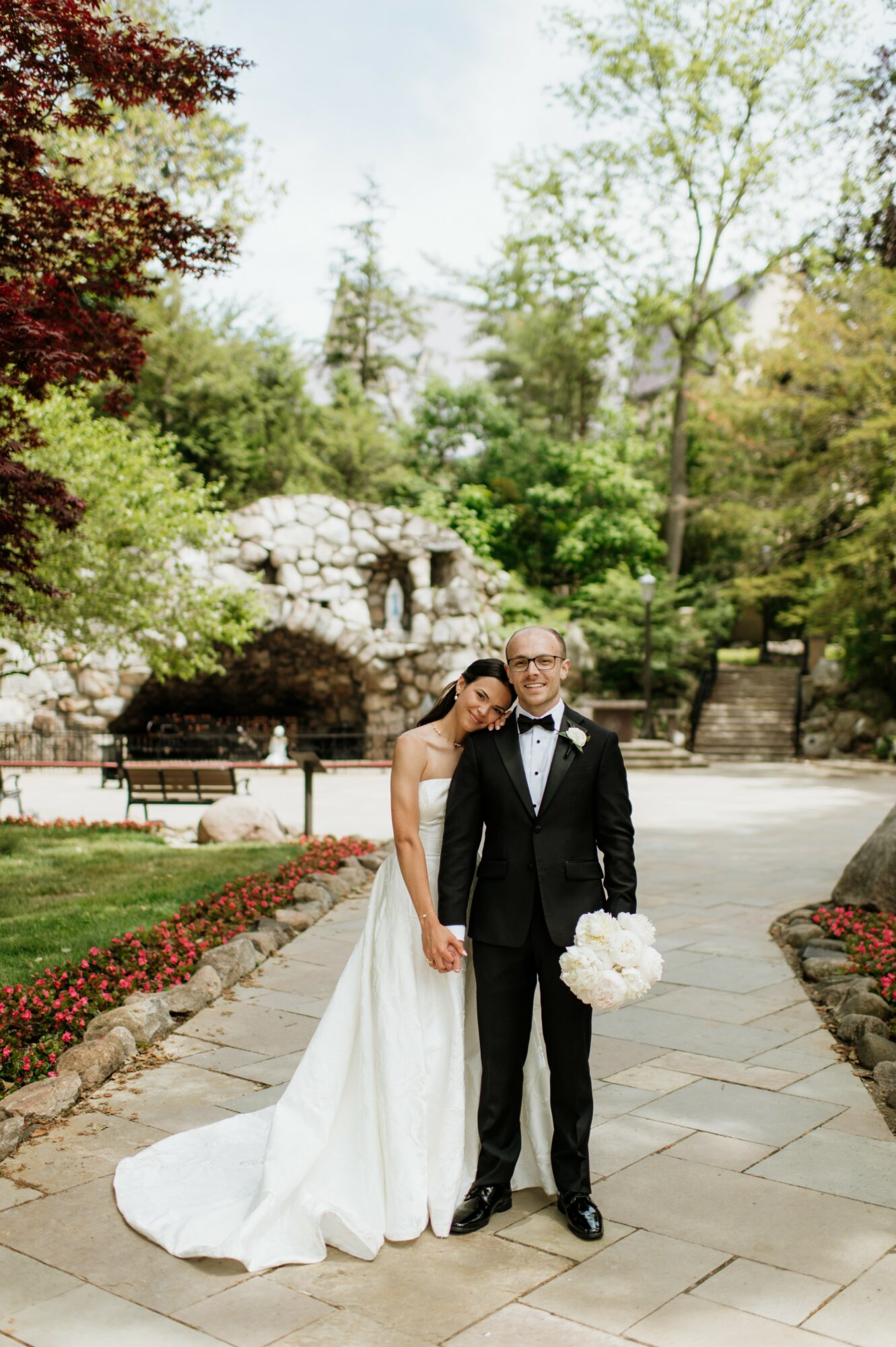 Bride and groom in wedding attire standing outdoors on a stone pathway, surrounded by trees and greenery, holding a bouquet.