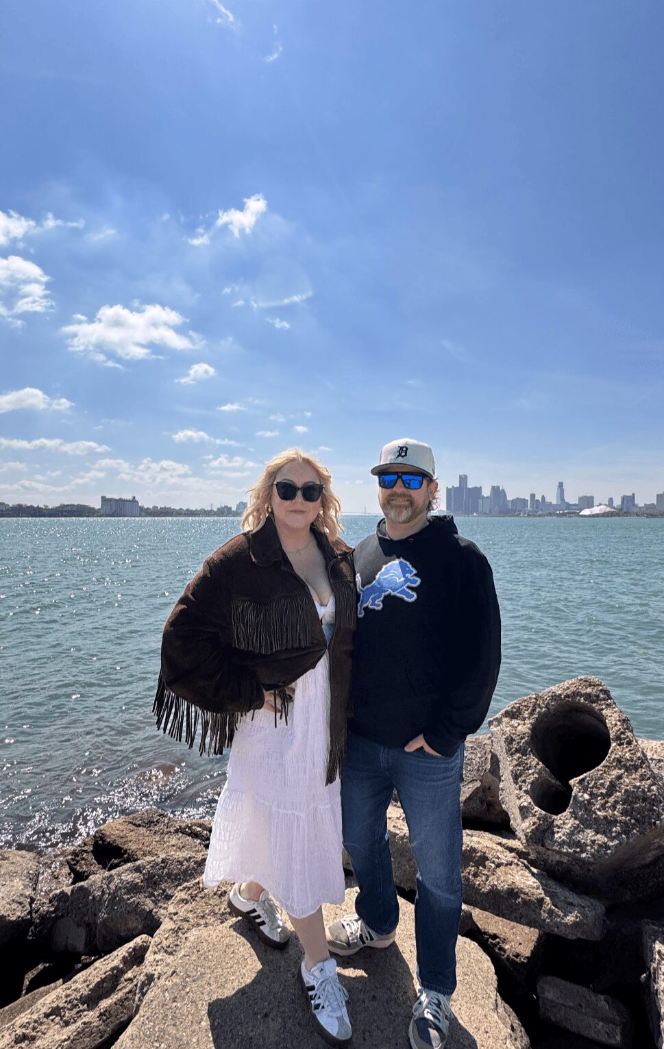 Two people stand on rocks by water with a city skyline in the background, under a partly cloudy sky.
