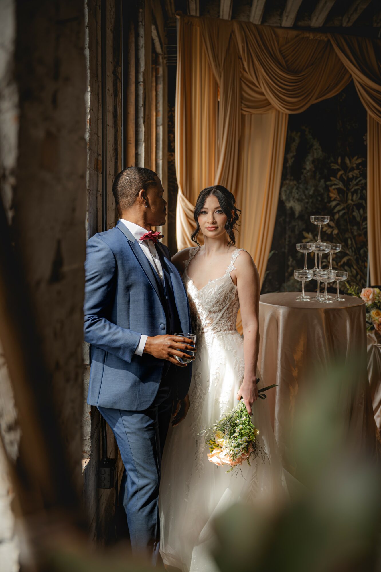 A bride in a white wedding dress holding a bouquet stands next to a groom in a blue suit indoors with curtains and a table with a champagne tower.
