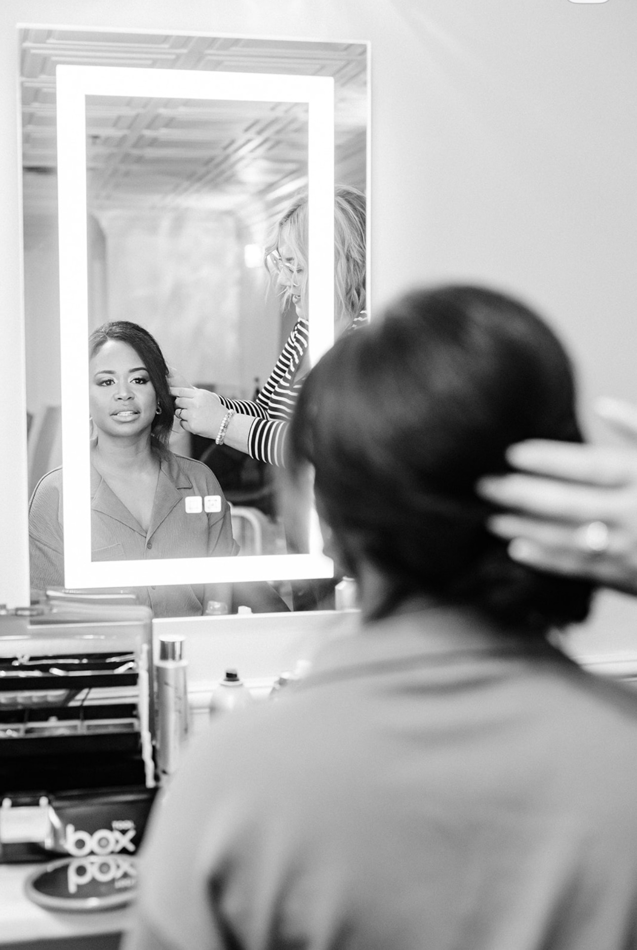Person with short hair looking at a mirror with a woman reflected in it, in a salon or similar setting.