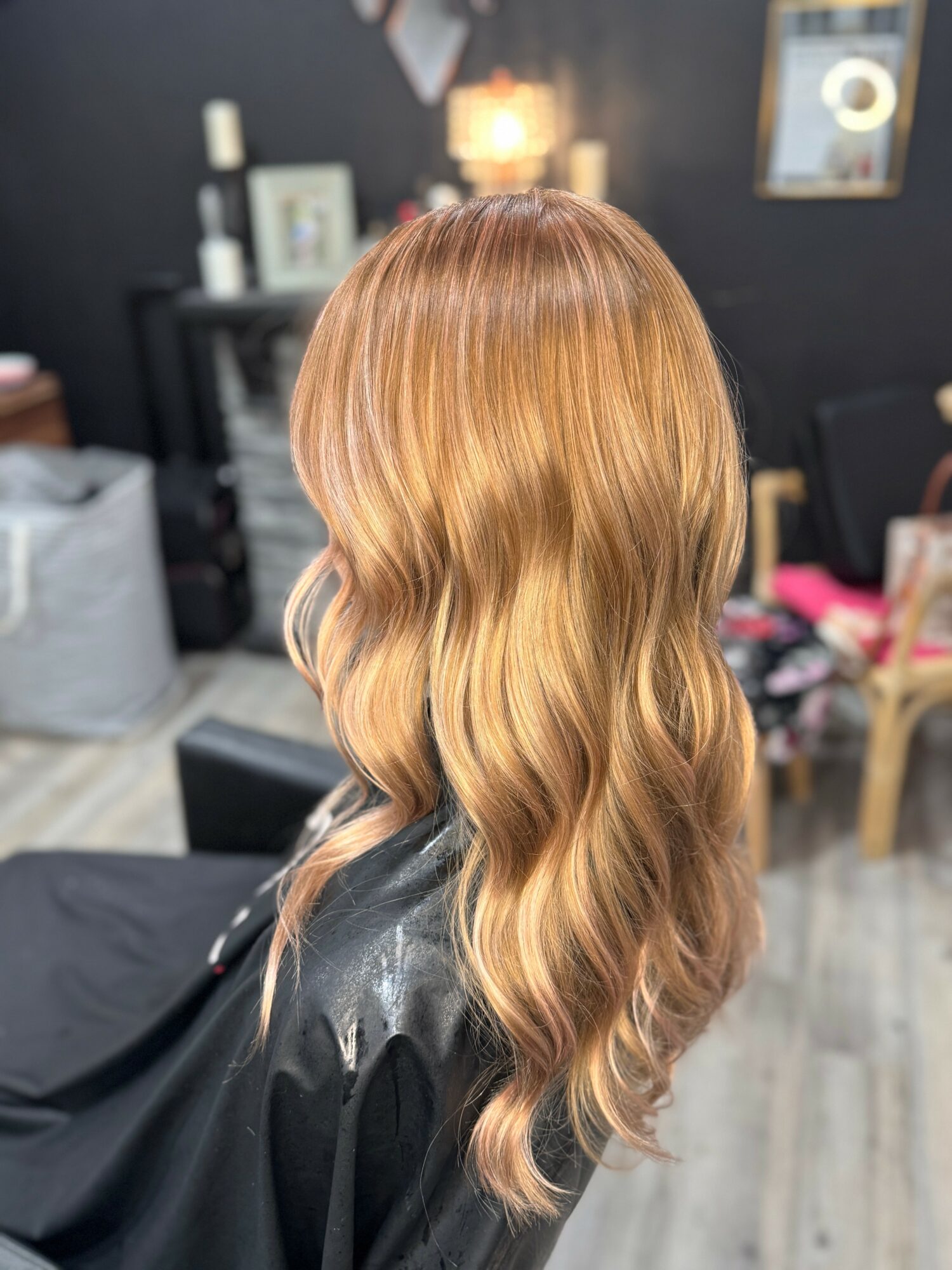 Back view of a woman with long, wavy, light brown hair in a salon, with blurred background of salon interior.