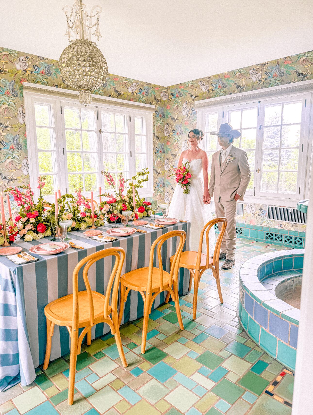 Bride and groom standing near windows with floral wallpaper, a decorated table with chairs, and a small pool in a bright room.