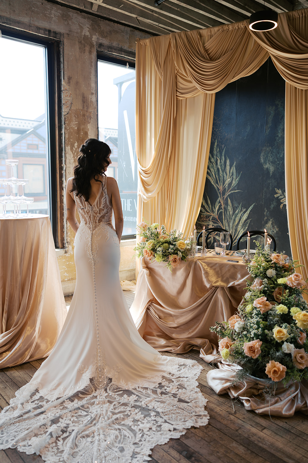 Bride in a white wedding dress stands indoors near large windows with natural light, decorated with draped curtains and floral arrangements.