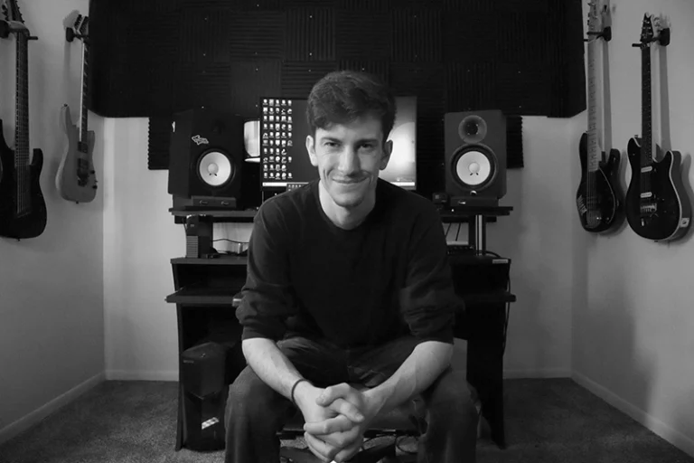 Young man sitting in a music studio with guitars on walls and speakers behind him, smiling, in black and white.