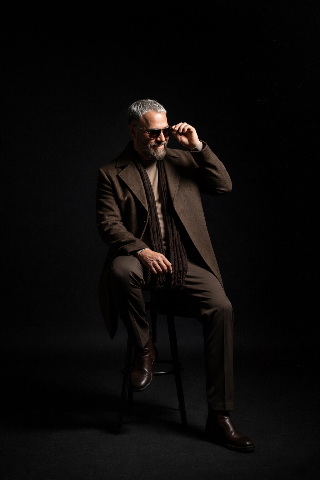 Man with gray hair and beard, wearing sunglasses, sitting on a stool, adjusting his glasses, in a dark setting.