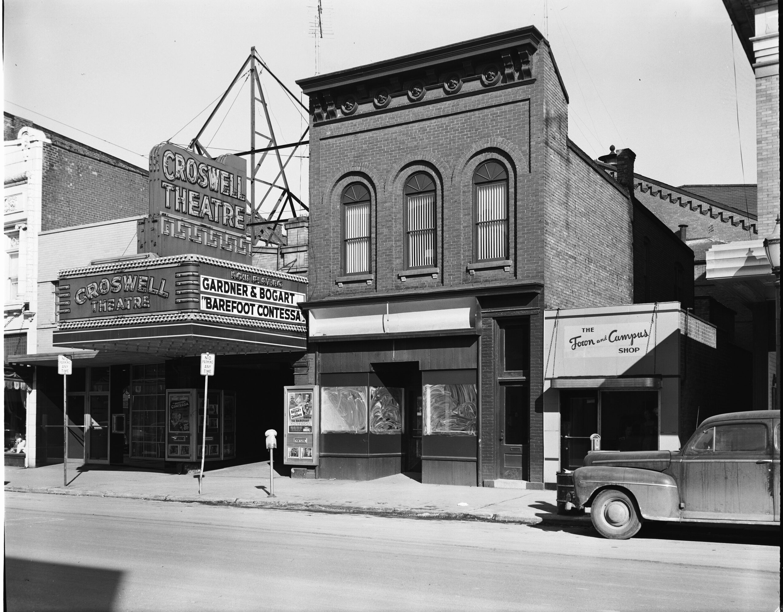 Street view of a brick building with a theater marquee, adjacent storefronts, and a parked vintage car.