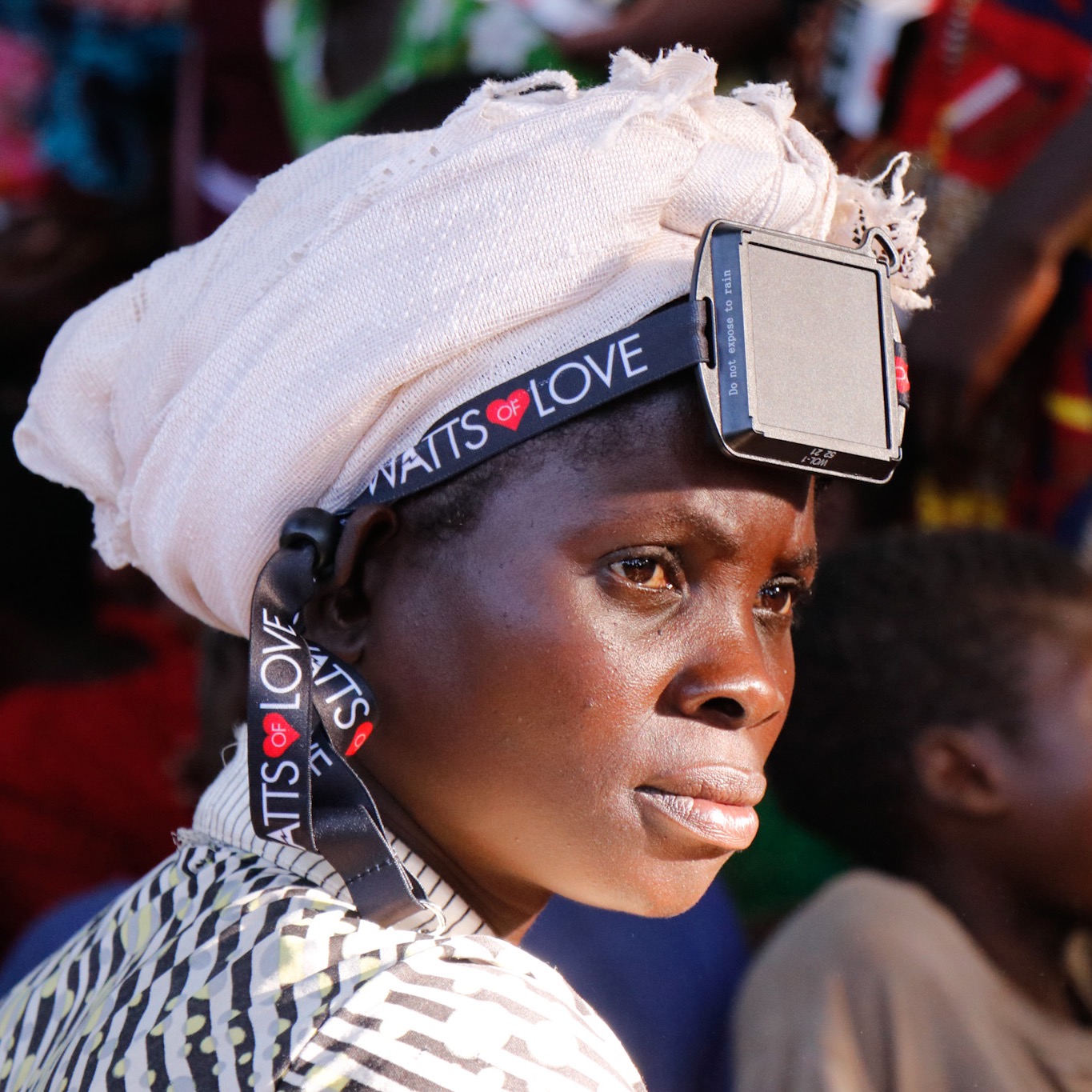Woman with head wrapped in white cloth, wearing a headlamp, looking to the side, in a crowded outdoor setting.
