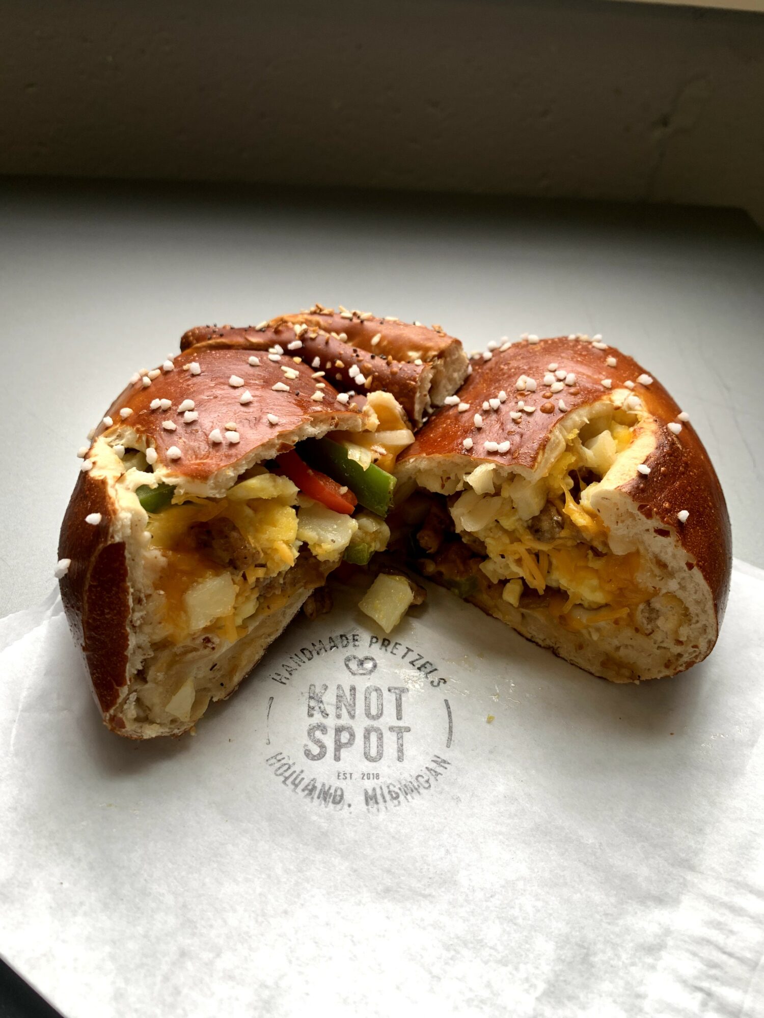 Broken pretzel bun filled with cheese, vegetables, and possibly meat, on white paper with a faint circular stamp, close-up.