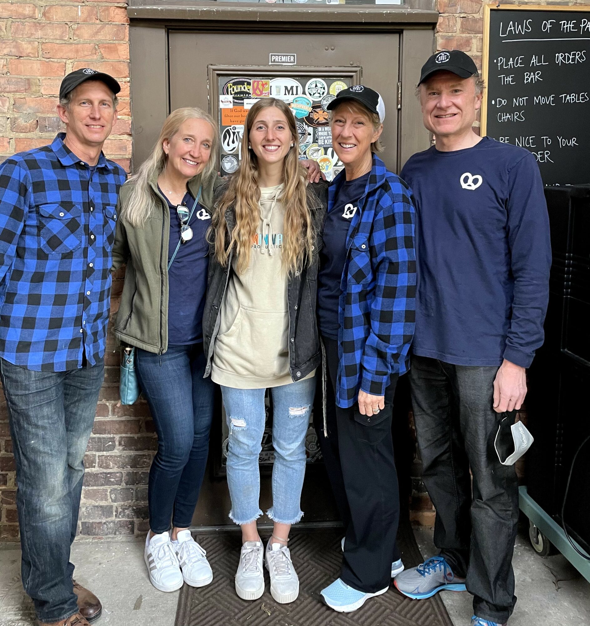 Five people stand outside a brick building, smiling, wearing casual clothing and hats, with a chalkboard and stickers behind them.